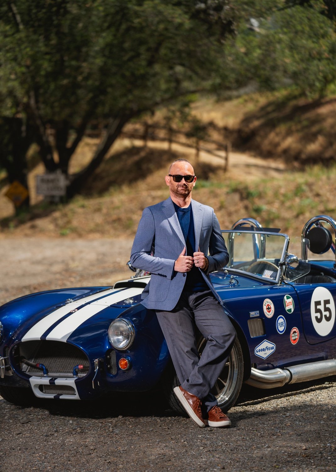 A man wearing sunglasses and a blazer leans against a vintage blue race car with racing stickers, standing on a gravel road with trees in the background.