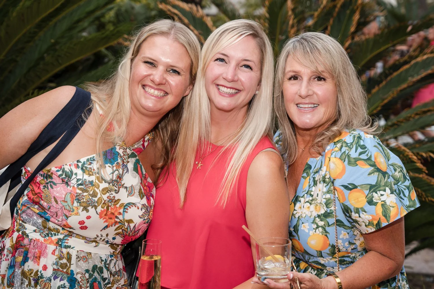 Three women smiling and standing together at an outdoor event, with tropical plants in the background. They are dressed in colorful summer clothing and holding beverages.