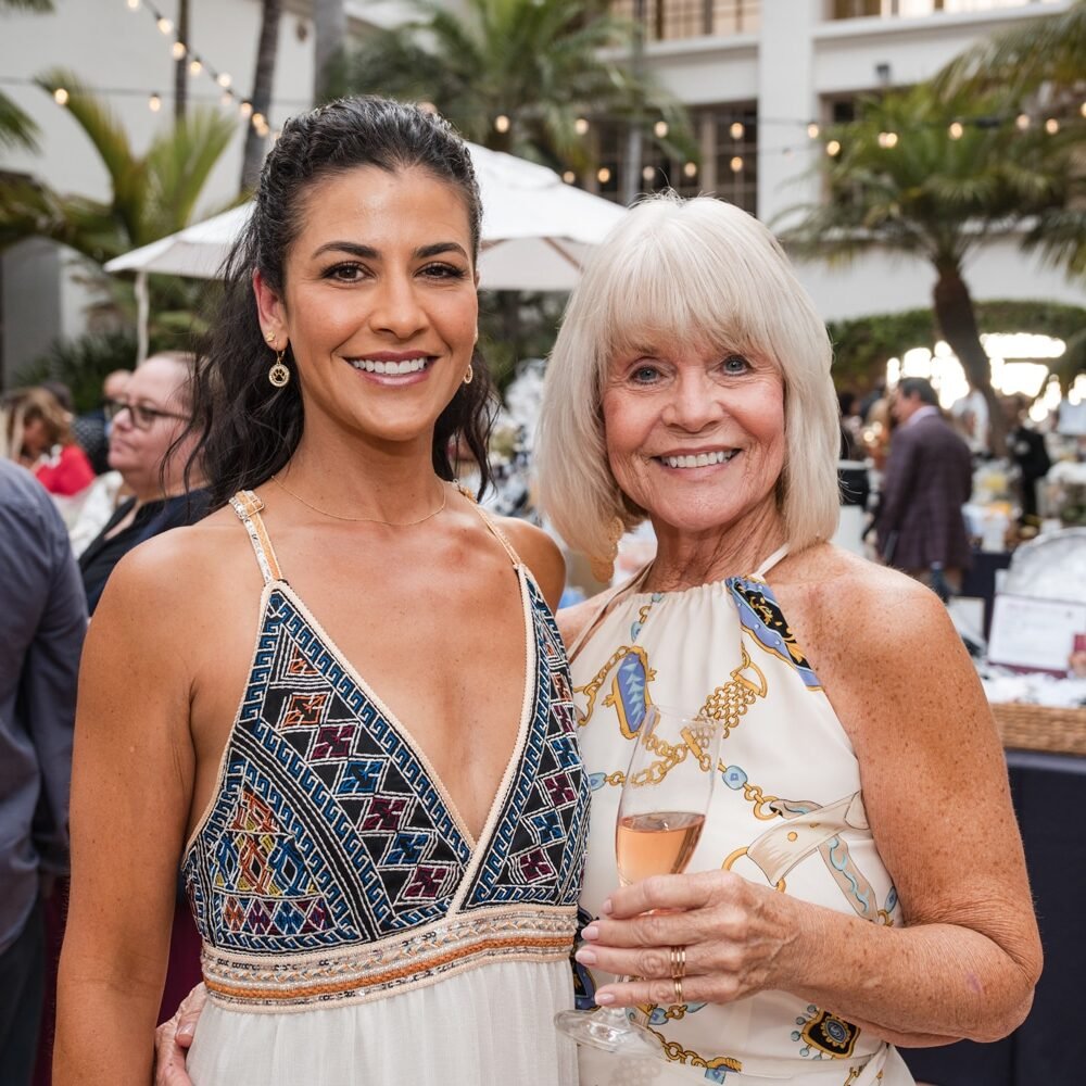 Two women smiling at an outdoor event, one holding a glass of rosé wine, with a decorated terrace and palm trees in the background.