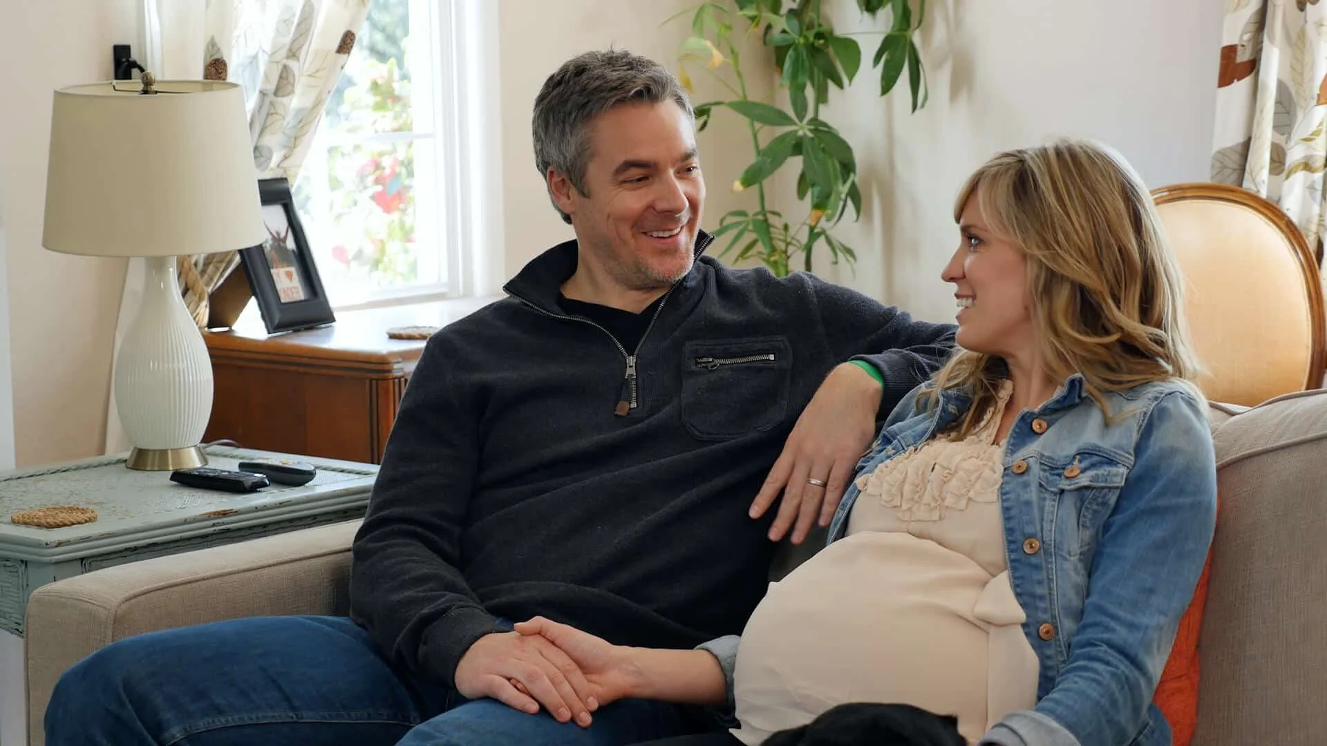 A man and woman sitting on a beige sofa in a living room, smiling and looking at each other, with the man wearing a black jacket and the woman wearing a denim jacket. There is a white lamp on a side table, a framed photograph, and a green potted plant in the background.