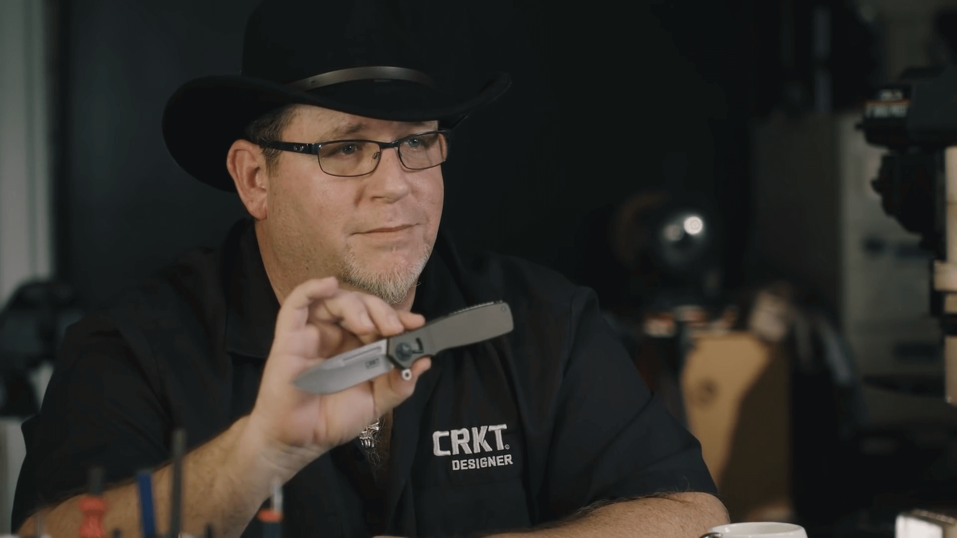 A man wearing glasses and a black cowboy hat, holding a multitool in his hand, in a workshop or garage setting.
