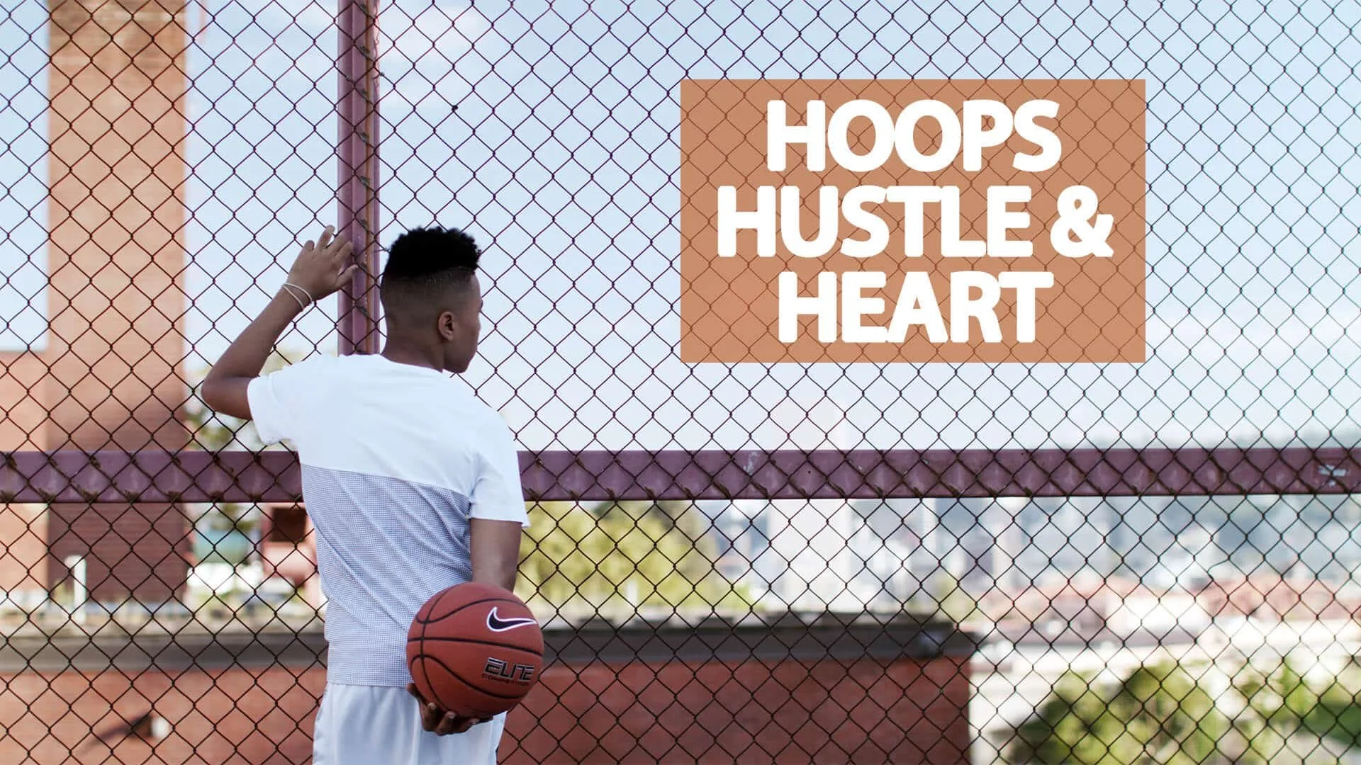 A young man in a white basketball uniform standing on a basketball court with a basketball in his hand, facing a chain-link fence, with a sign that reads 'Hoops Hustle & Heart' attached to the fence.