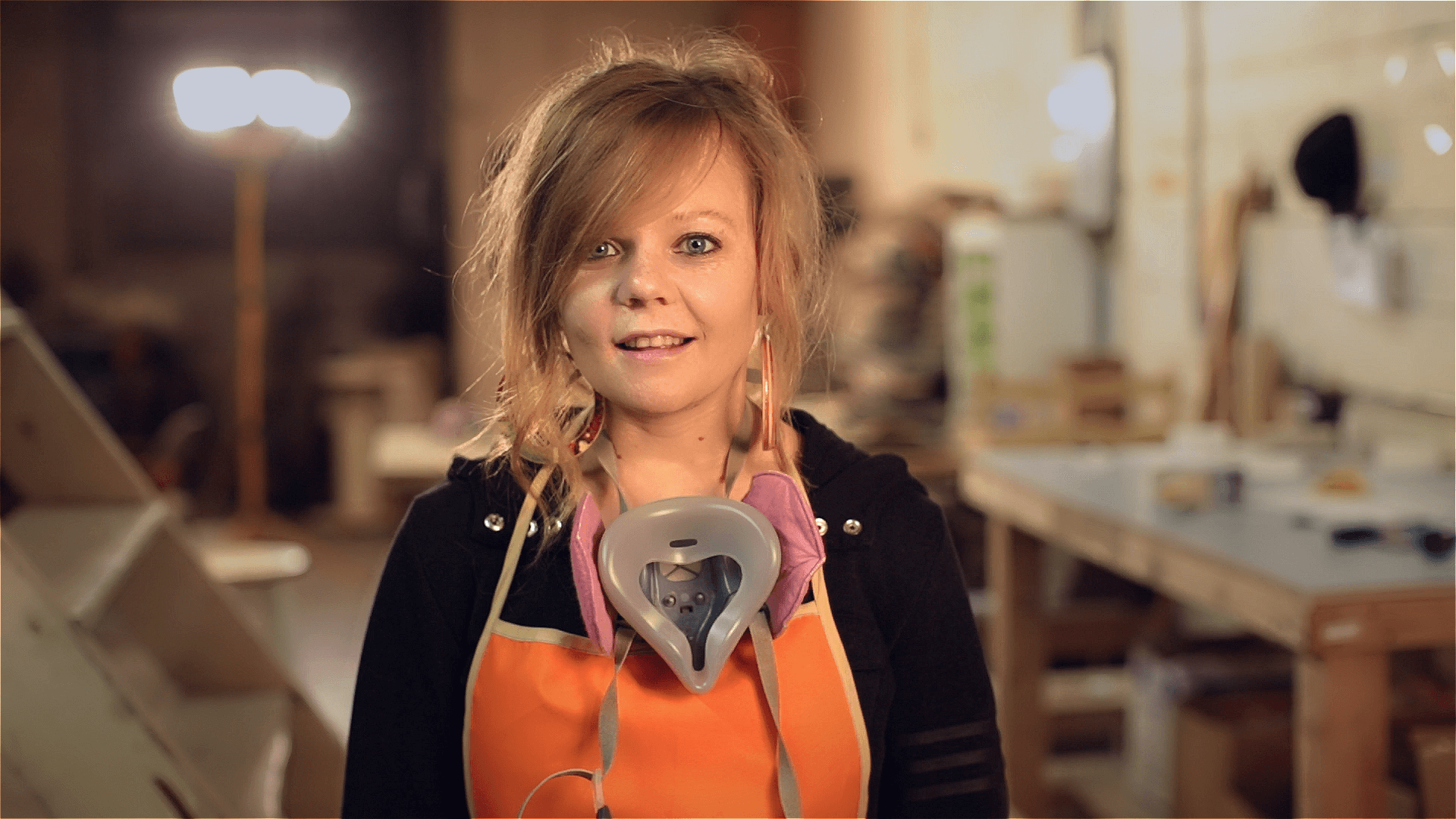 Woman with messy hair wearing an orange apron and a respirator mask around her neck, standing in a workshop or studio.