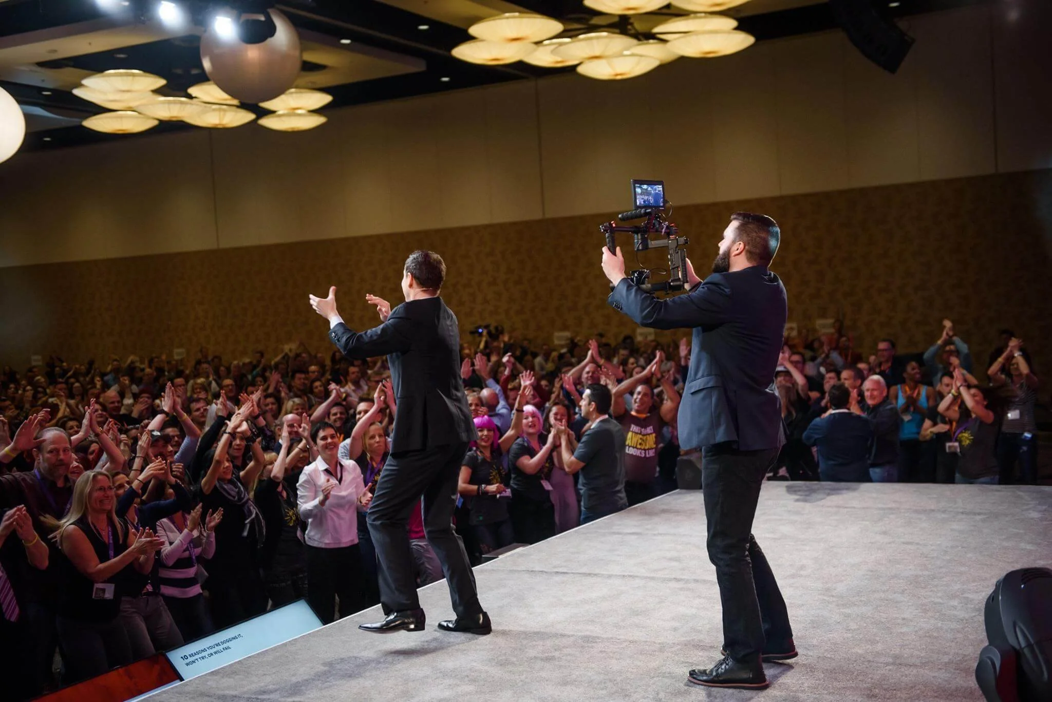 A speaker on stage with a microphone and camera facing him, standing in front of a lively audience at a conference or event in a large room with decorative ceiling lights.
