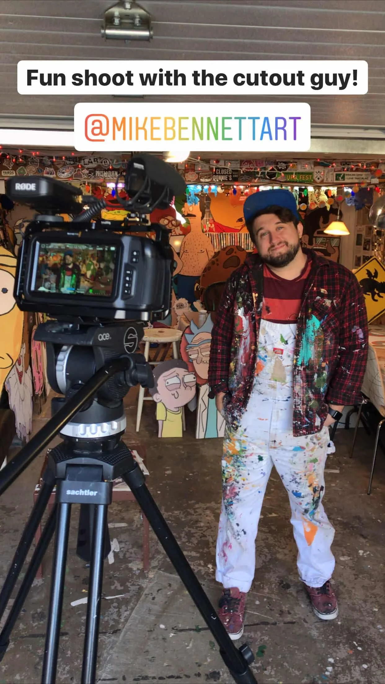 A man standing in front of cartoon character cutouts in a colorful art studio, wearing paint-splattered clothes and a blue cap, with a camera on a tripod recording him.
