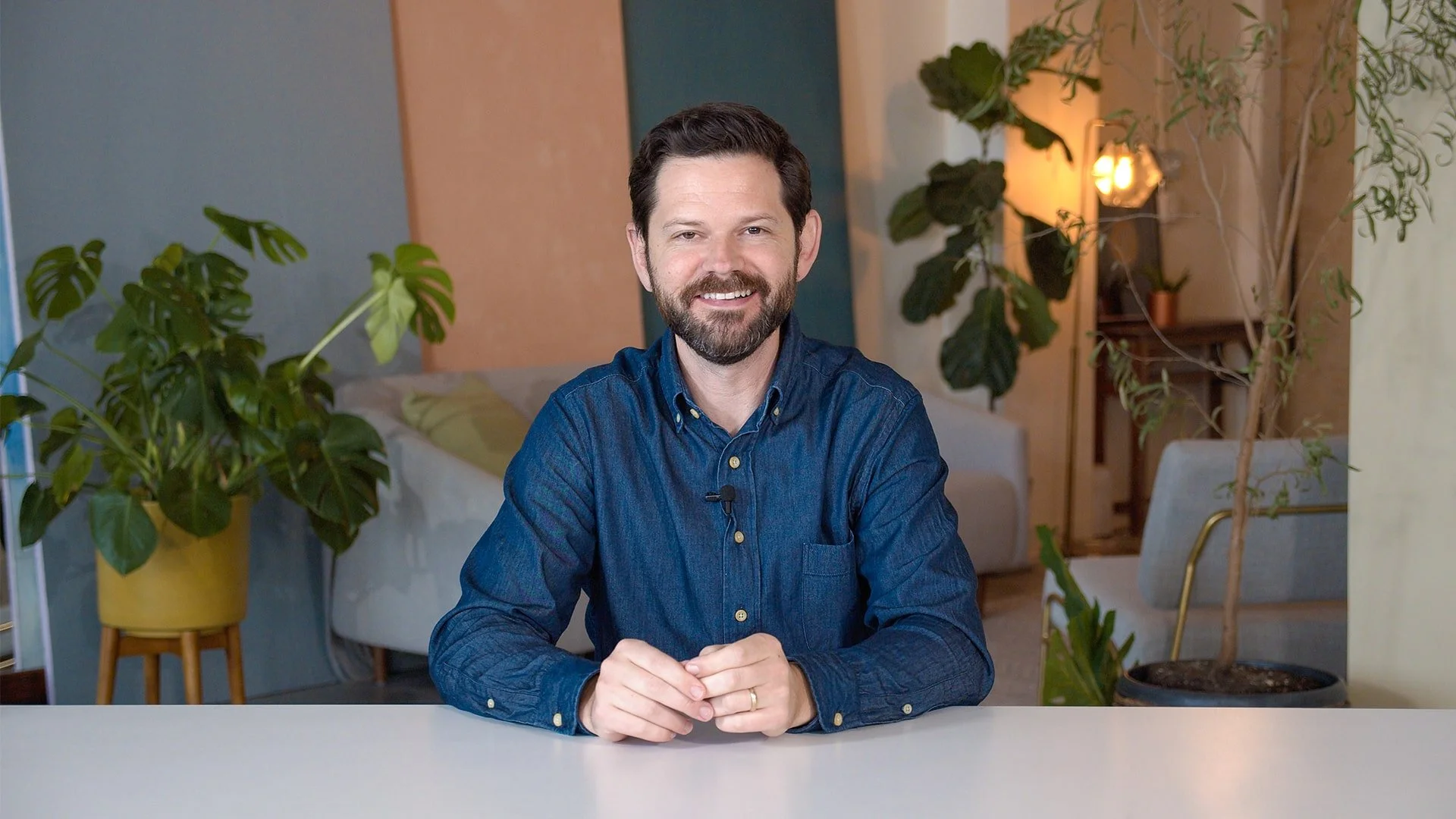 Smiling man with dark hair and beard, wearing a blue denim shirt, sitting at a table in a room with plants and warm lighting.