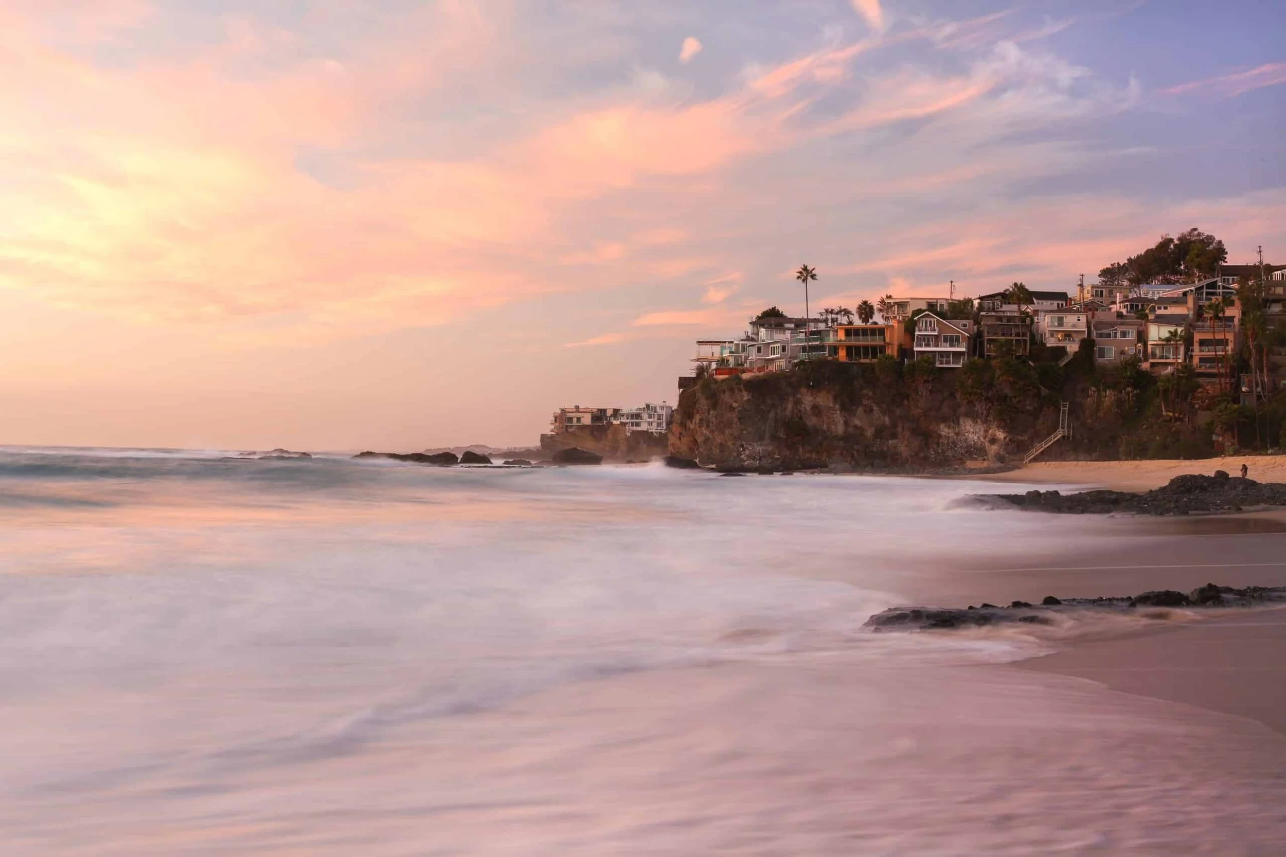 Beach at sunset with waves crashing on the shore, houses built on a rocky cliff, and palm trees against a pink and purple sky.