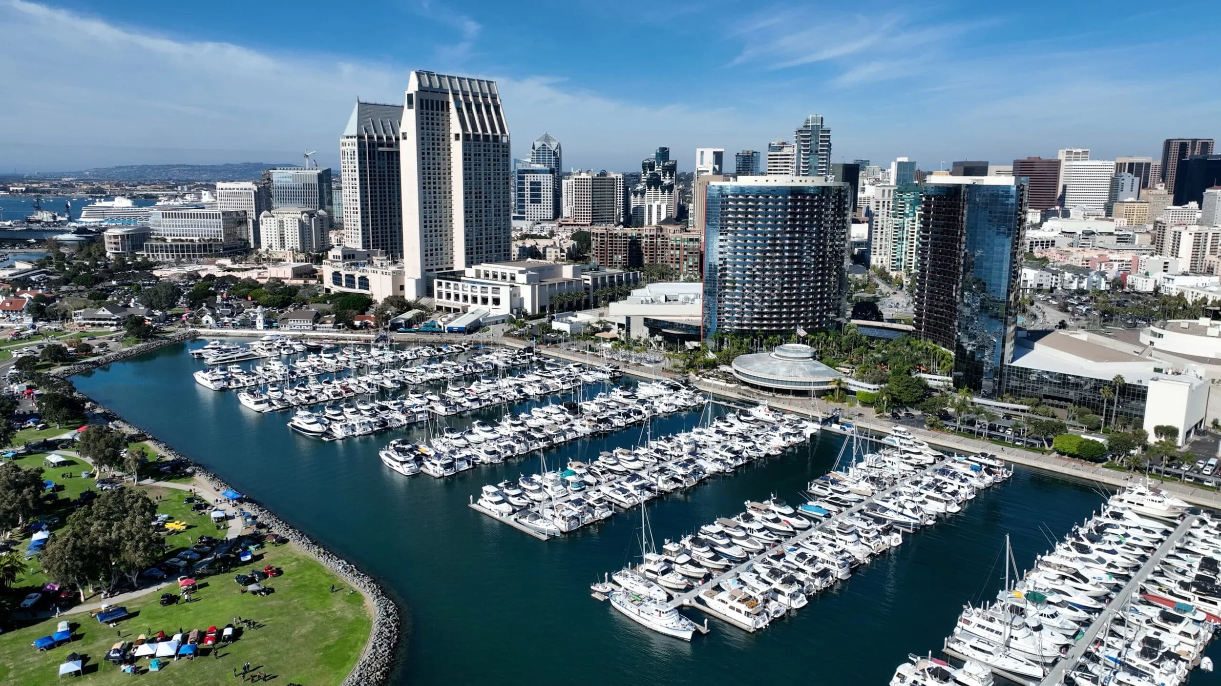 Aerial view of a city skyline with modern high-rise buildings and a marina filled with yachts, next to a park with tents and people.