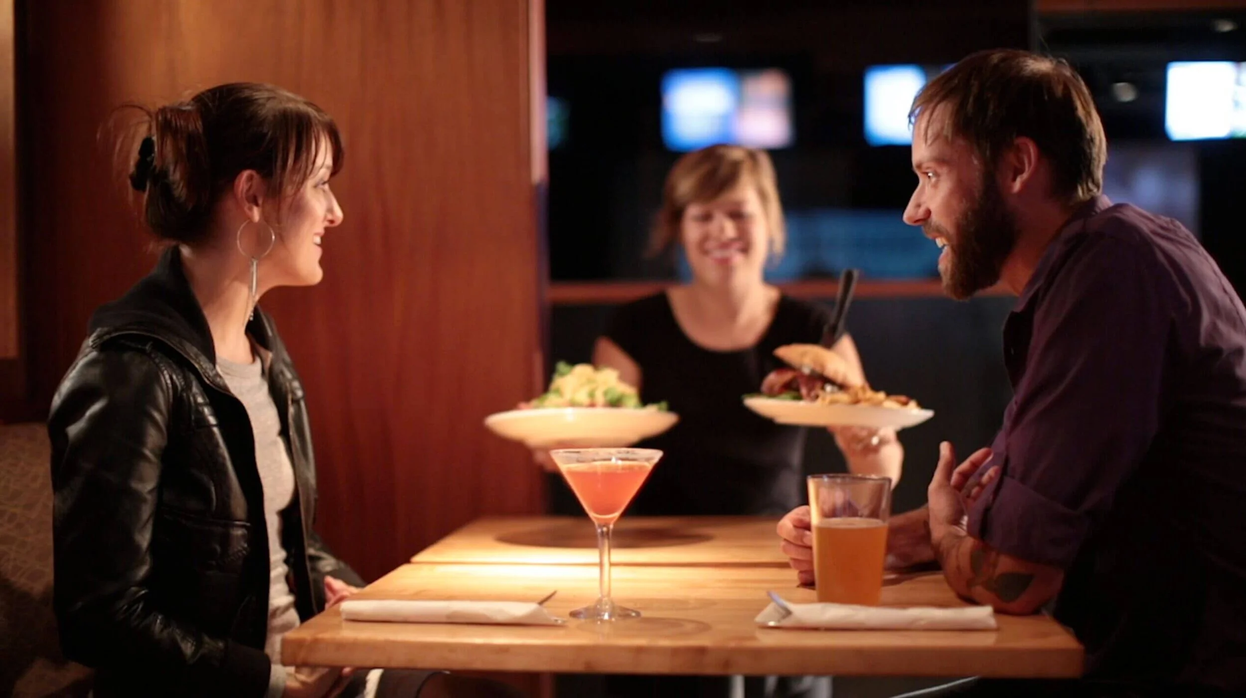 A man and woman are talking at a bar, with a waitress serving food in the background, and drinks on the table.