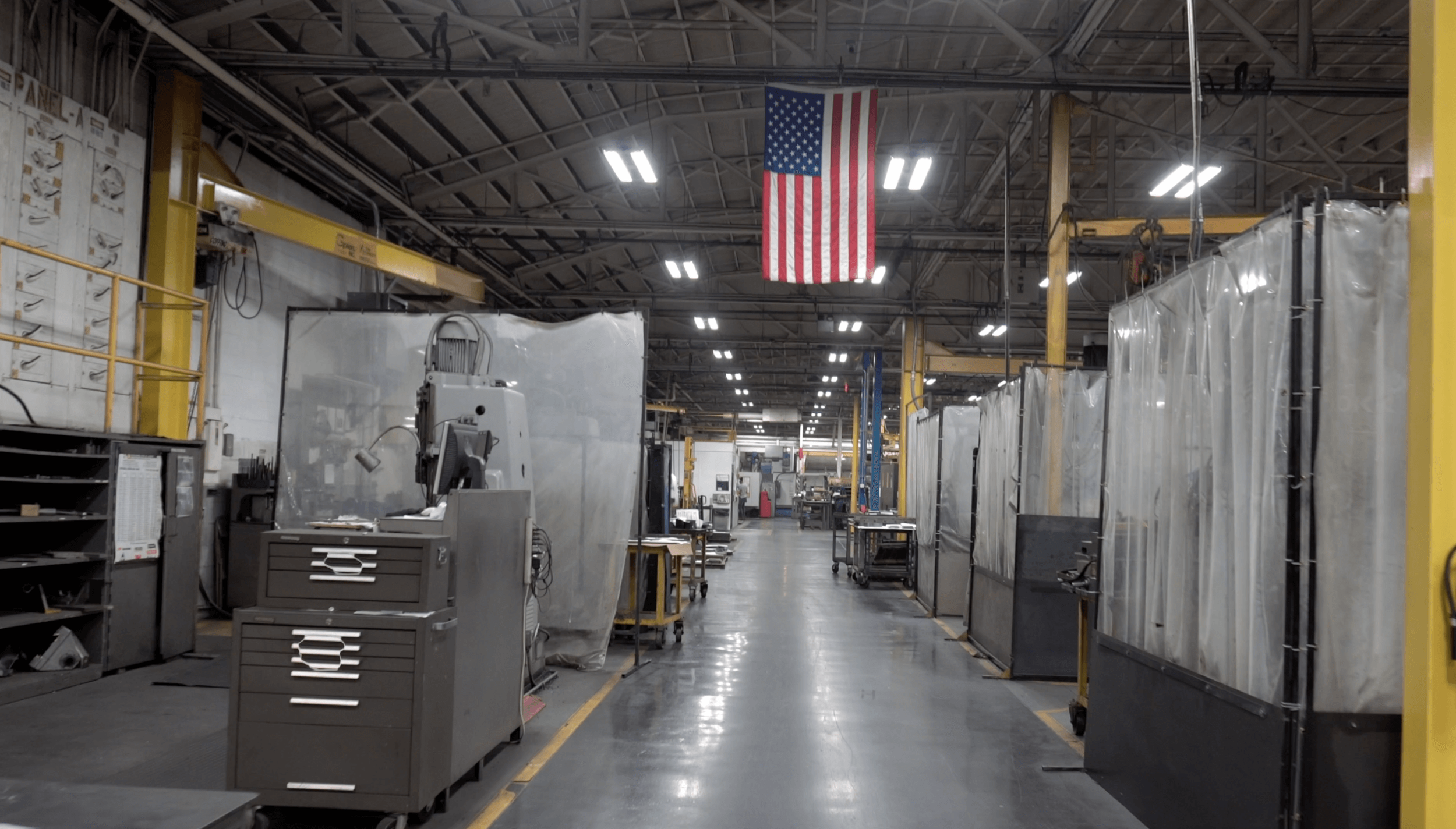 Interior of an industrial manufacturing facility with metal workstations, plastic curtains, machinery, and an American flag hanging from the ceiling.