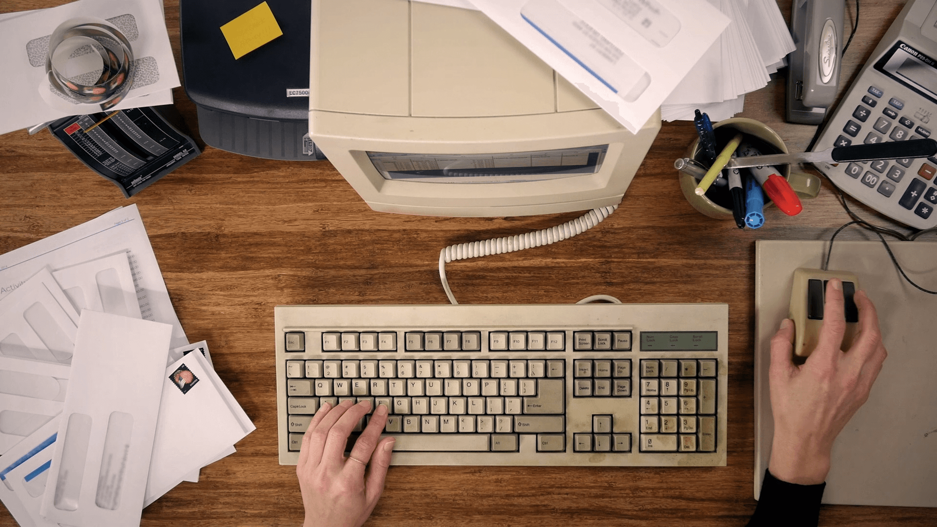 A cluttered desk with an old beige keyboard, a beige mouse, a computer monitor, scattered papers, a cup filled with pens and markers, a calculator, a desktop phone, and a person’s hand on the mouse.