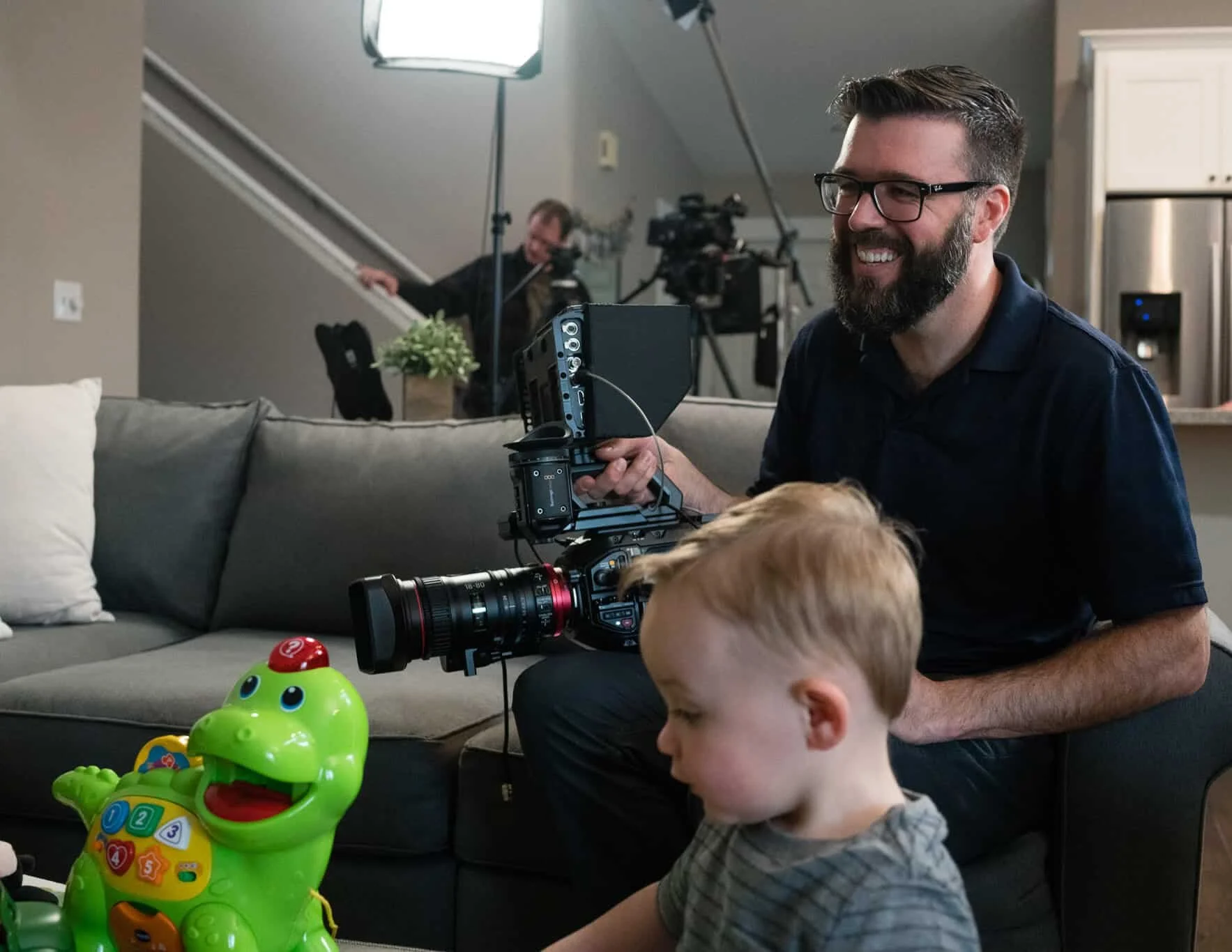 A man with glasses and a beard is sitting on a couch holding a professional camera, smiling at two young children who are playing with colorful plastic toys on the floor. In the background, a person is operating filming equipment inside a home setting.