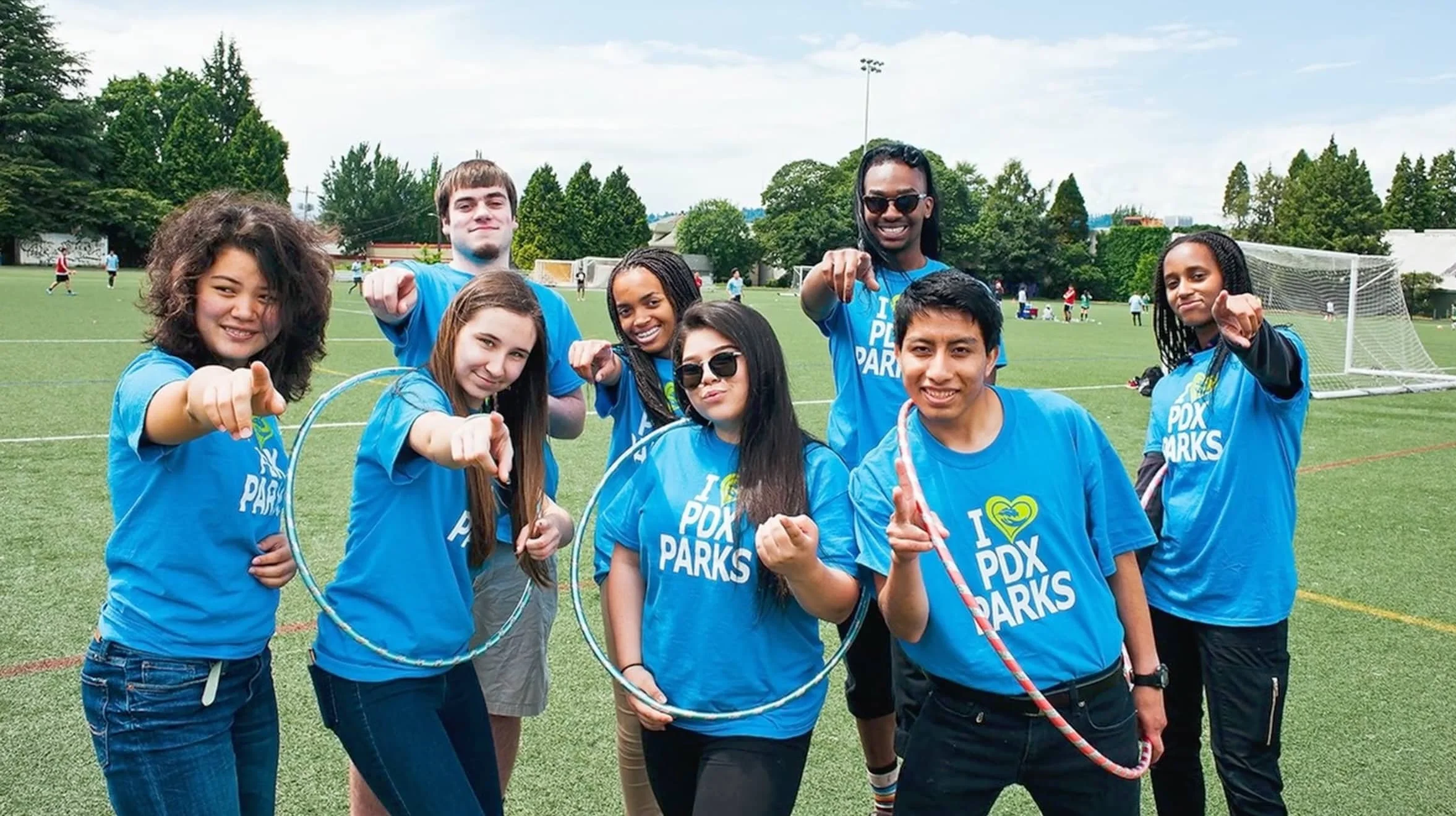 Group of young people wearing matching blue T-shirts with 'I ❤️ PDX Parks' logo, posing and pointing towards the camera on a grassy field during a sunny day.