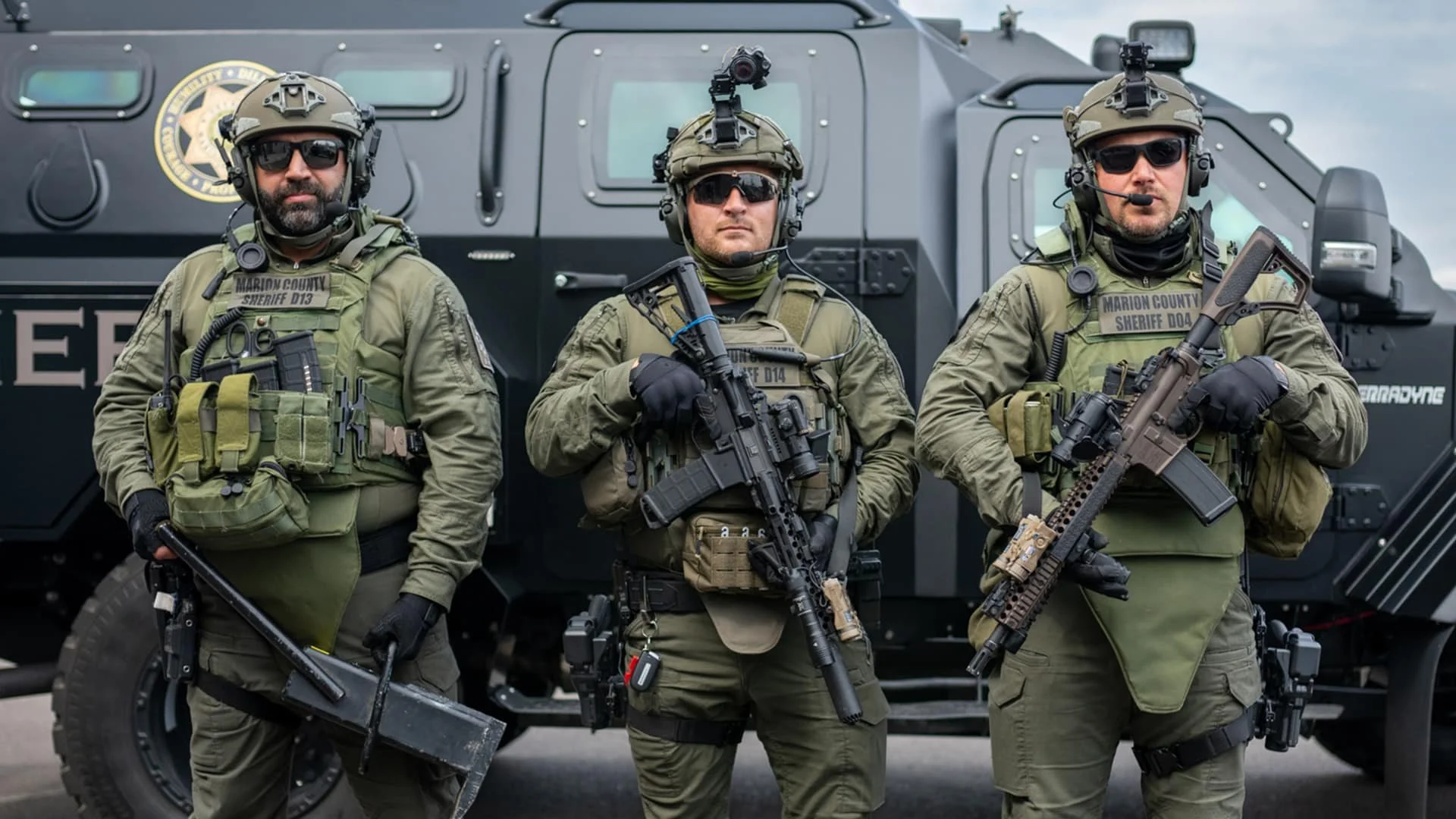 Three armed police officers in tactical gear standing in front of a black armored vehicle.