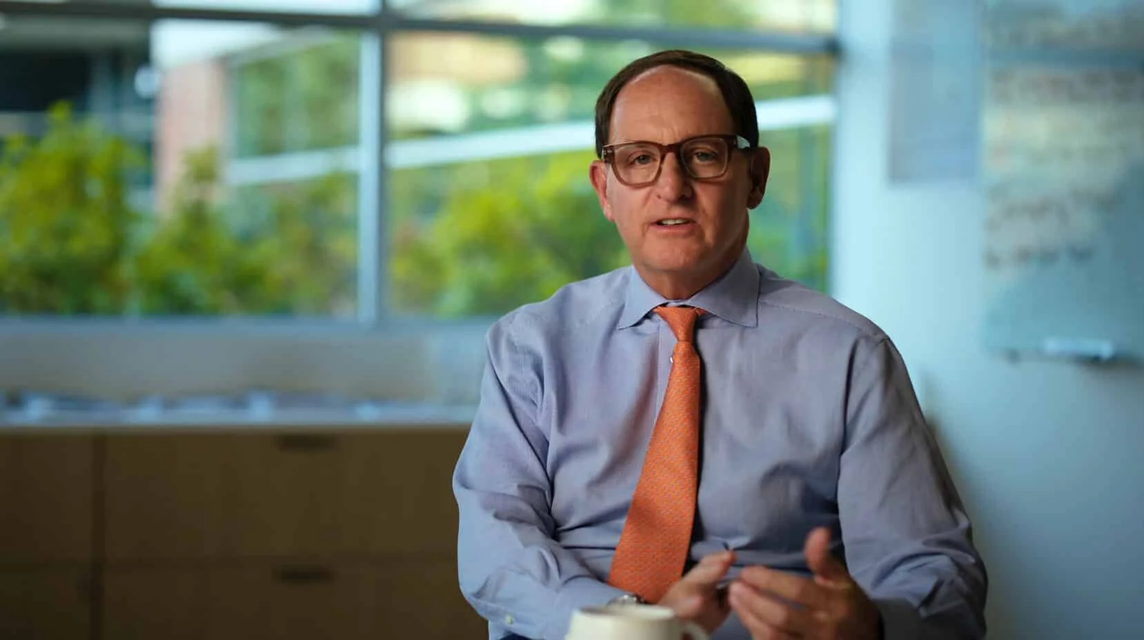 A man wearing glasses, a light blue dress shirt, and an orange tie sitting in an office with large windows showing trees outside.