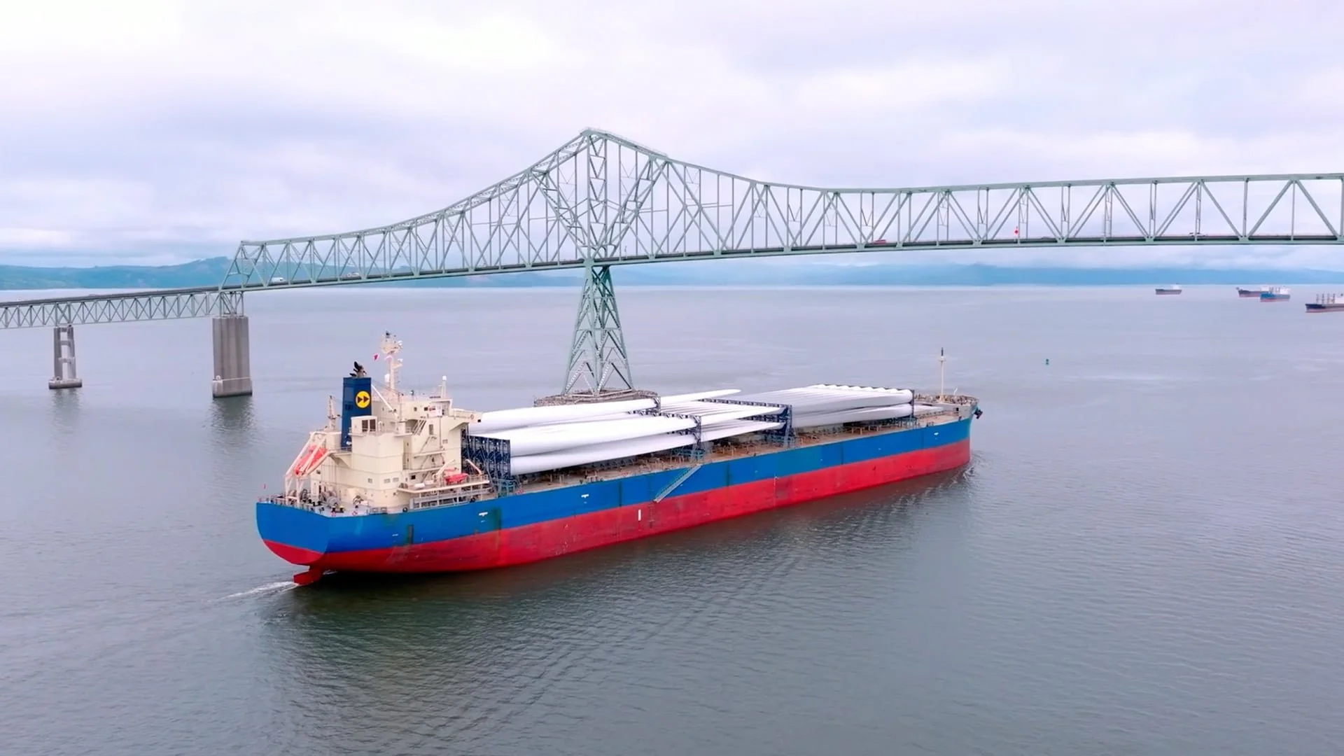 A large cargo ship carrying wind turbine components sailing under a bridge over calm water with several cargo ships in the distance, cloudy sky, and hills in the background.