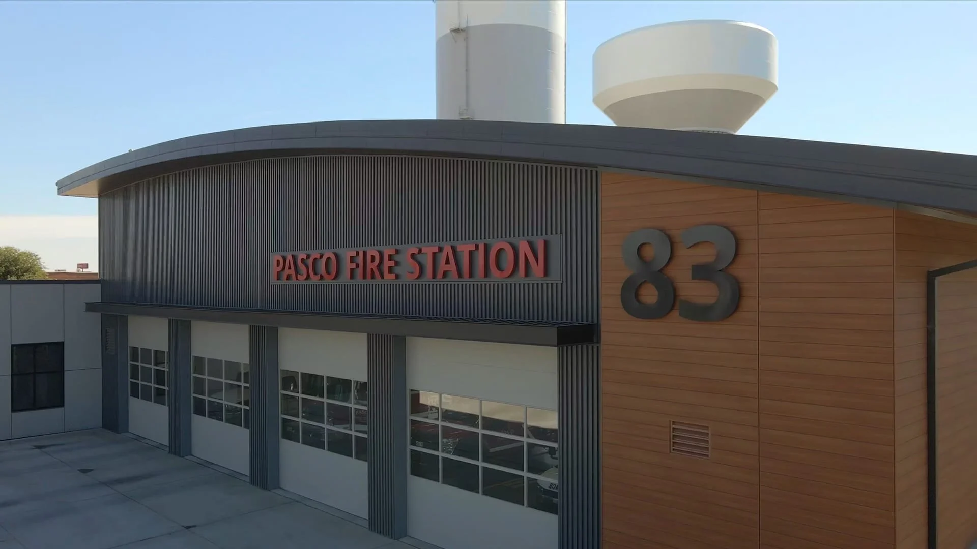 Exterior view of Pasco Fire Station 83 building with modern design, gray metal siding, wooden accents, large windows, and a prominent sign with red letters and gray numbers.