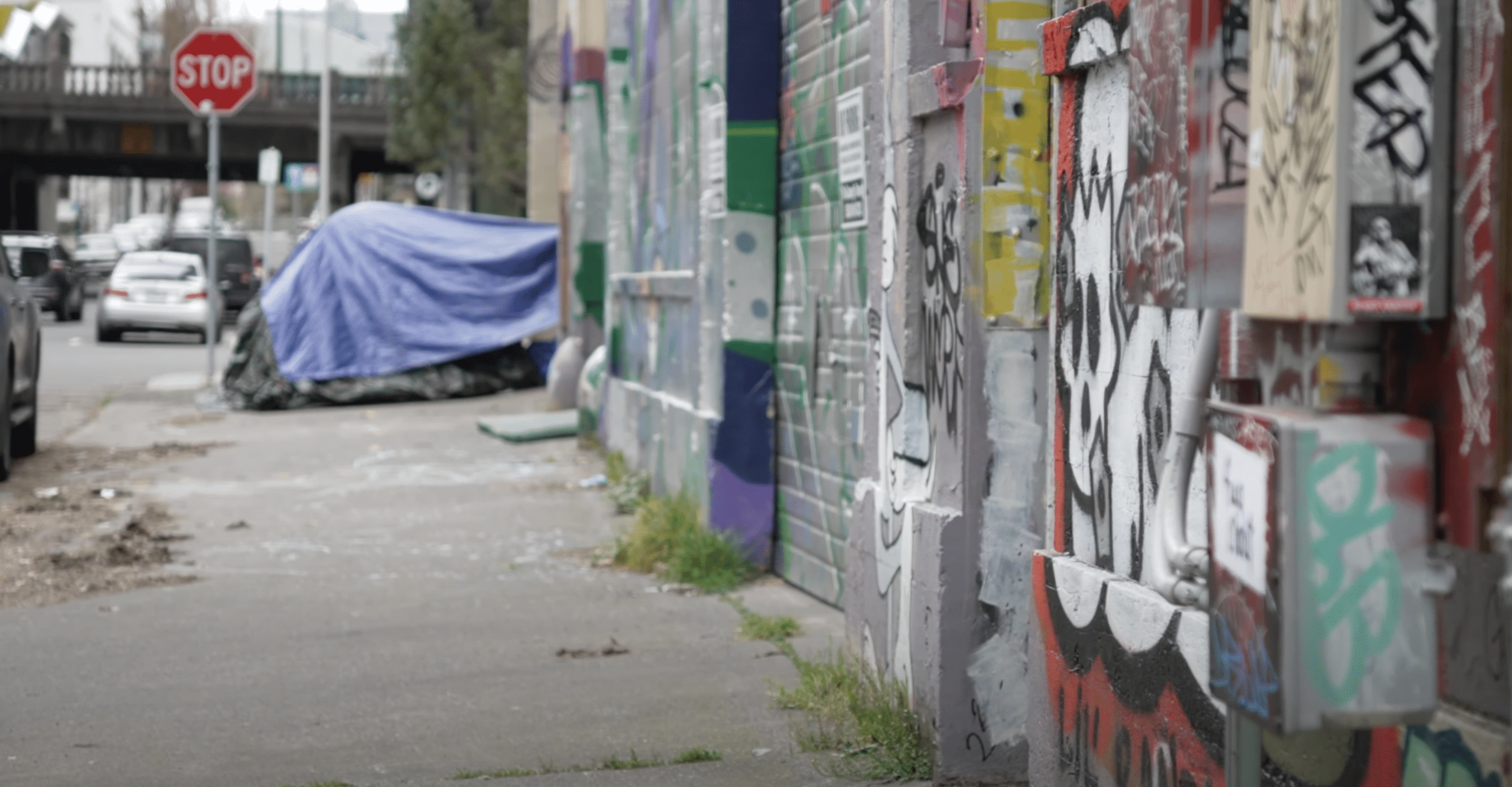A graffiti-covered metal fence runs along a city sidewalk next to a street with parked and moving cars. There is a blue tarp covering an object on the sidewalk. In the background, there is a stop sign and an overpass bridge.