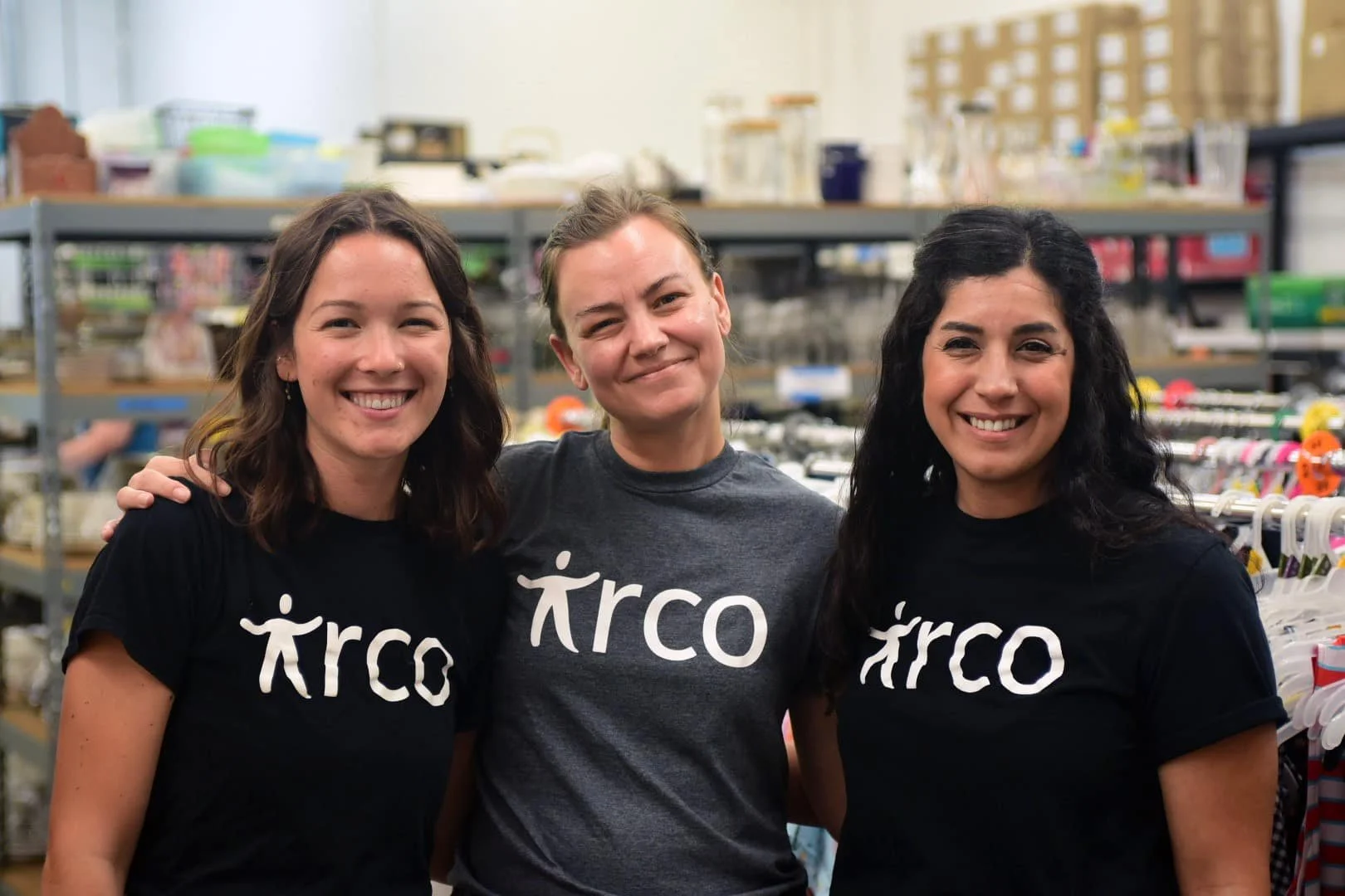 Three women smiling and standing close together in a store, wearing matching ARCO shirts. Shelves of supplies are visible in the background.