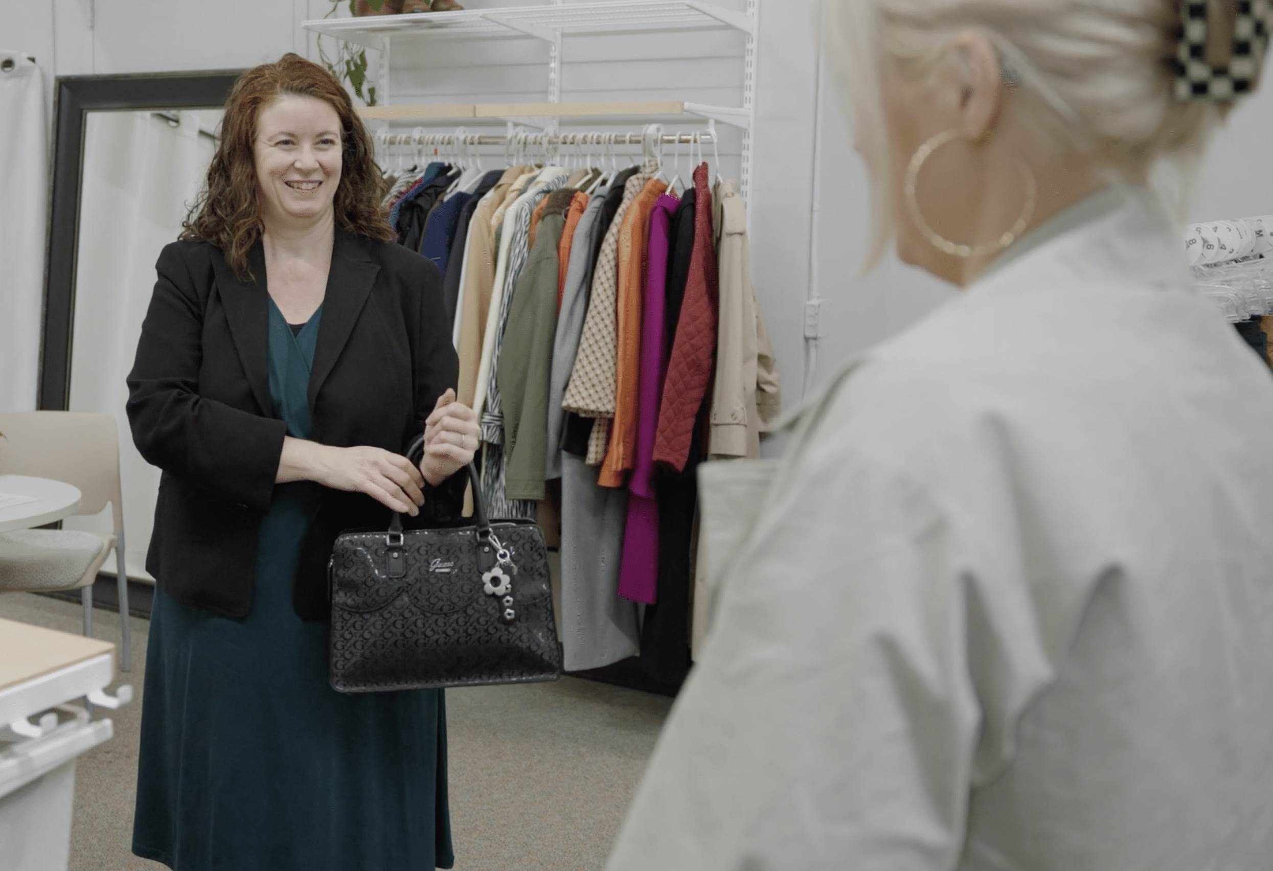Two women are inside a clothing store, shaking hands. One woman has red hair and wearing a blazer, the other has blonde hair tied back. Clothing is displayed on racks in the background.