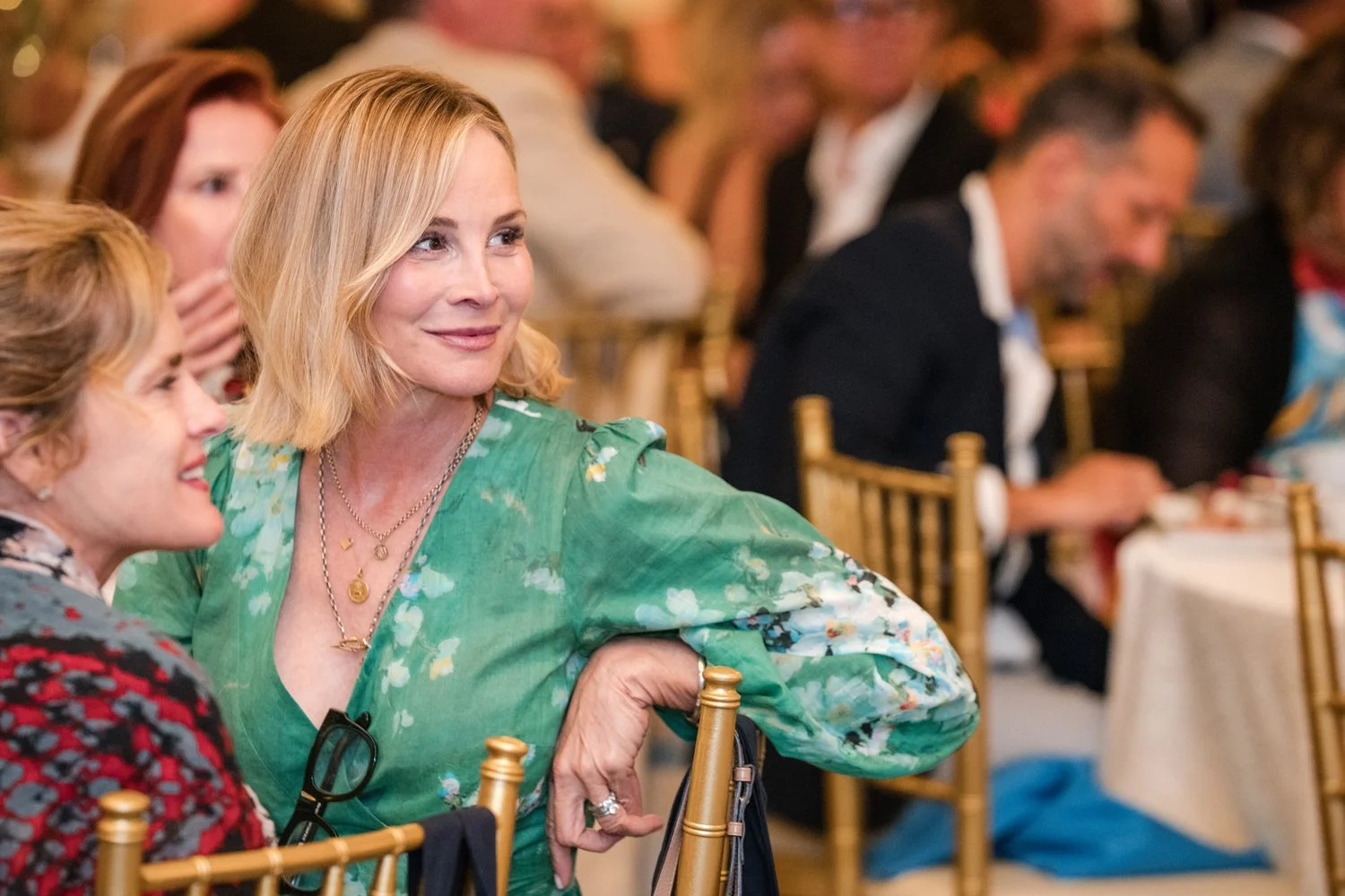 A woman with shoulder-length blonde hair, smiling and dressed in a green floral dress, sitting at a formal event. She has layered gold necklaces and a ring on her right hand, which is resting on the back of a gold chair.