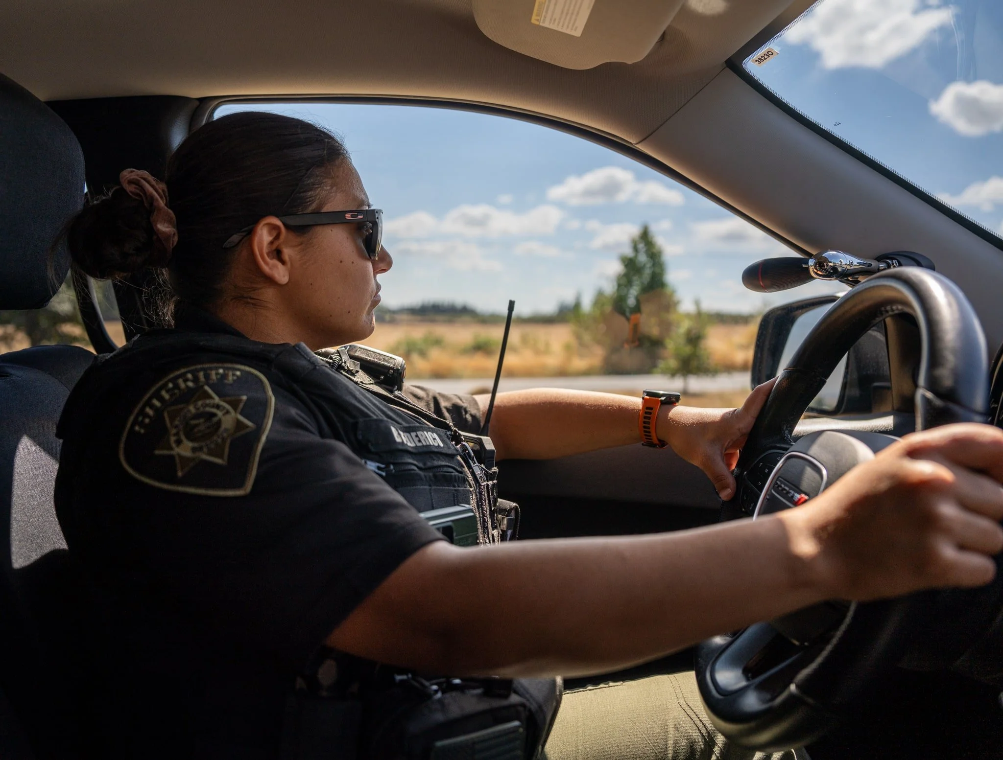 A woman in a police uniform wearing sunglasses, sitting in the passenger seat of a vehicle, looking out the window at a rural landscape under a partly cloudy sky.