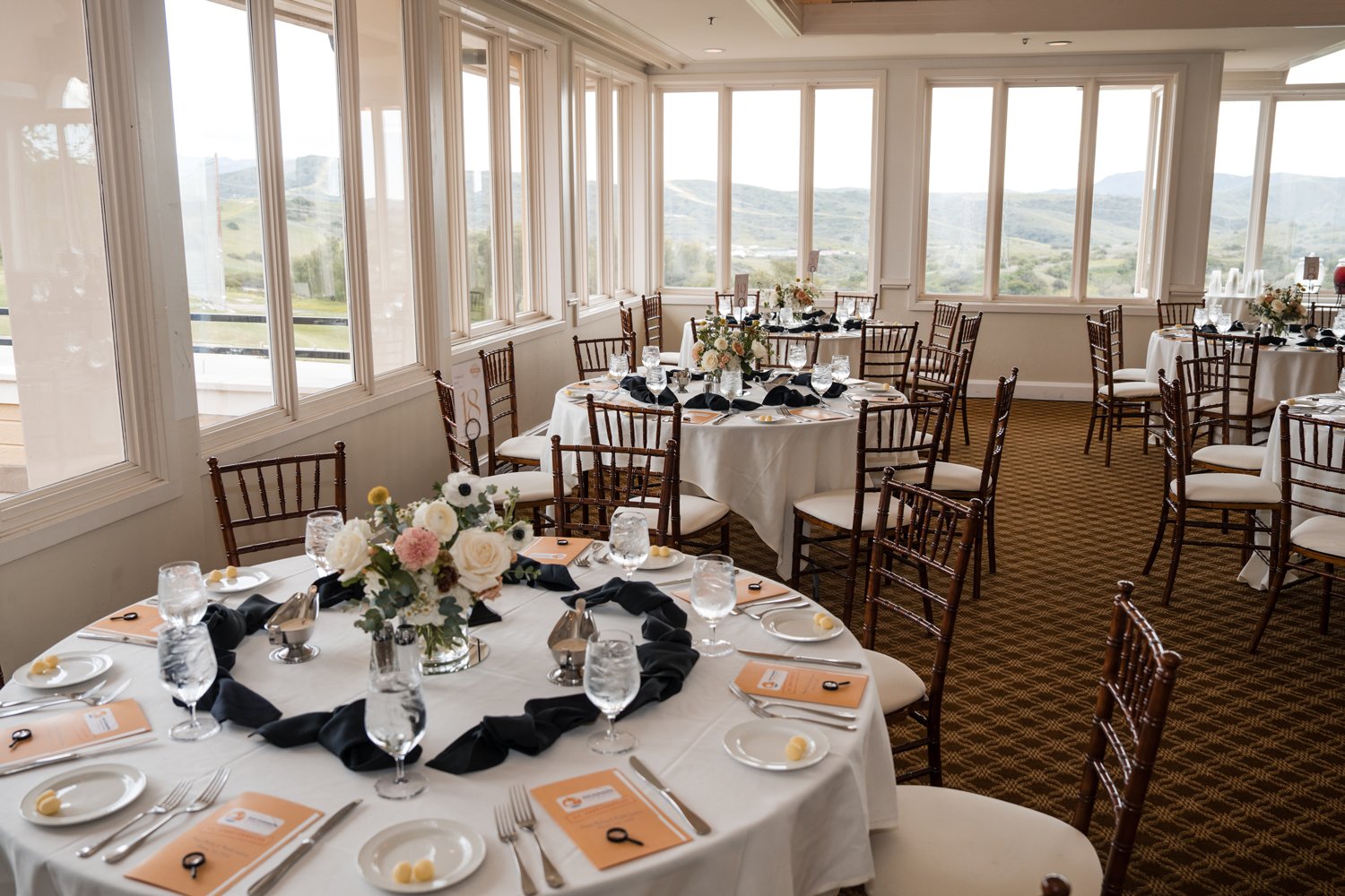 A banquet room decorated for a formal event with round tables covered with white tablecloths, black napkins, glassware, plates, and floral centerpieces, surrounded by wooden chairs, with large windows and a scenic view outside.
