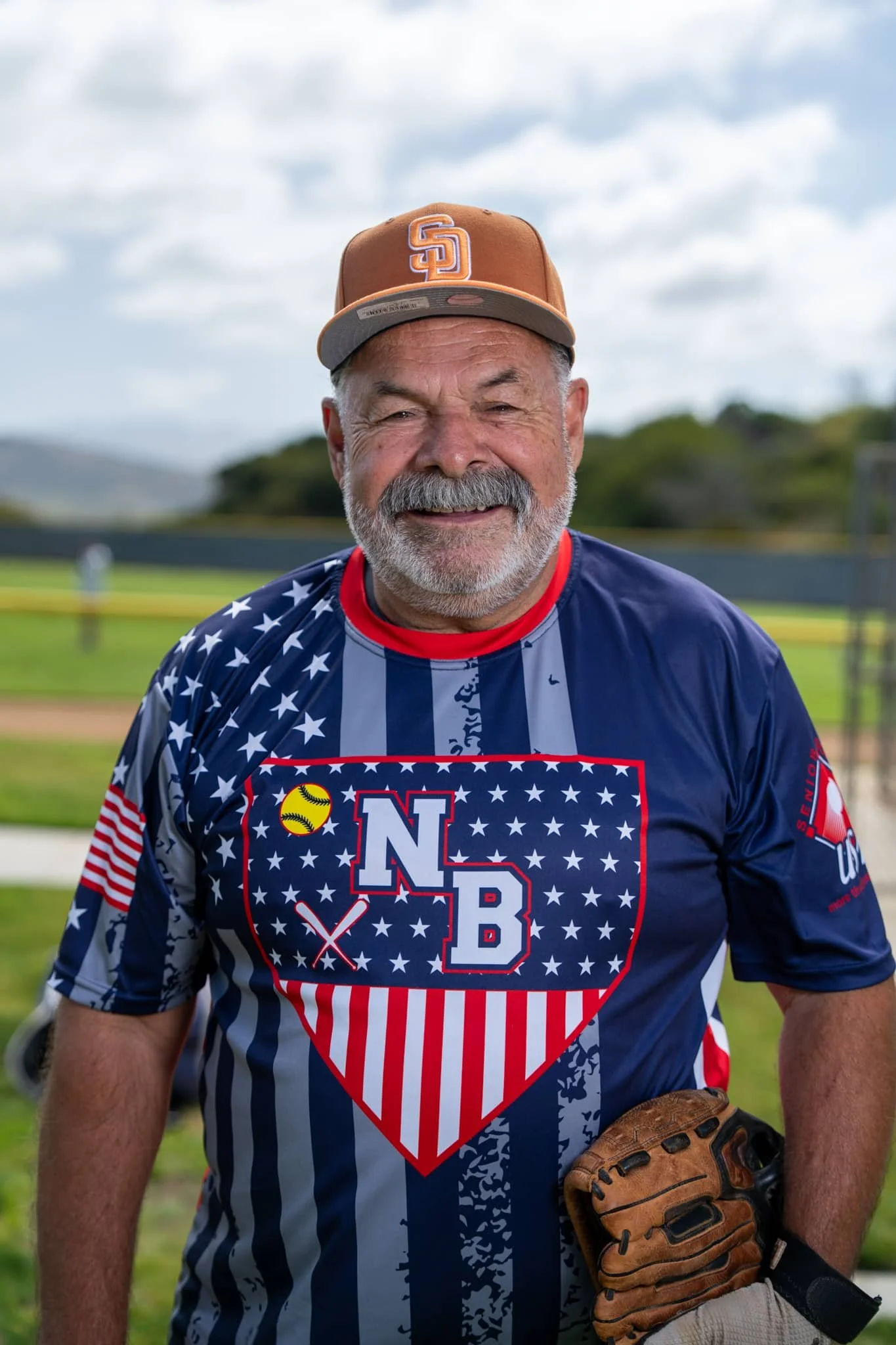 A smiling man wearing a baseball cap and a patriotic baseball jersey with stars and stripes, standing on a baseball field with a glove in his left hand.