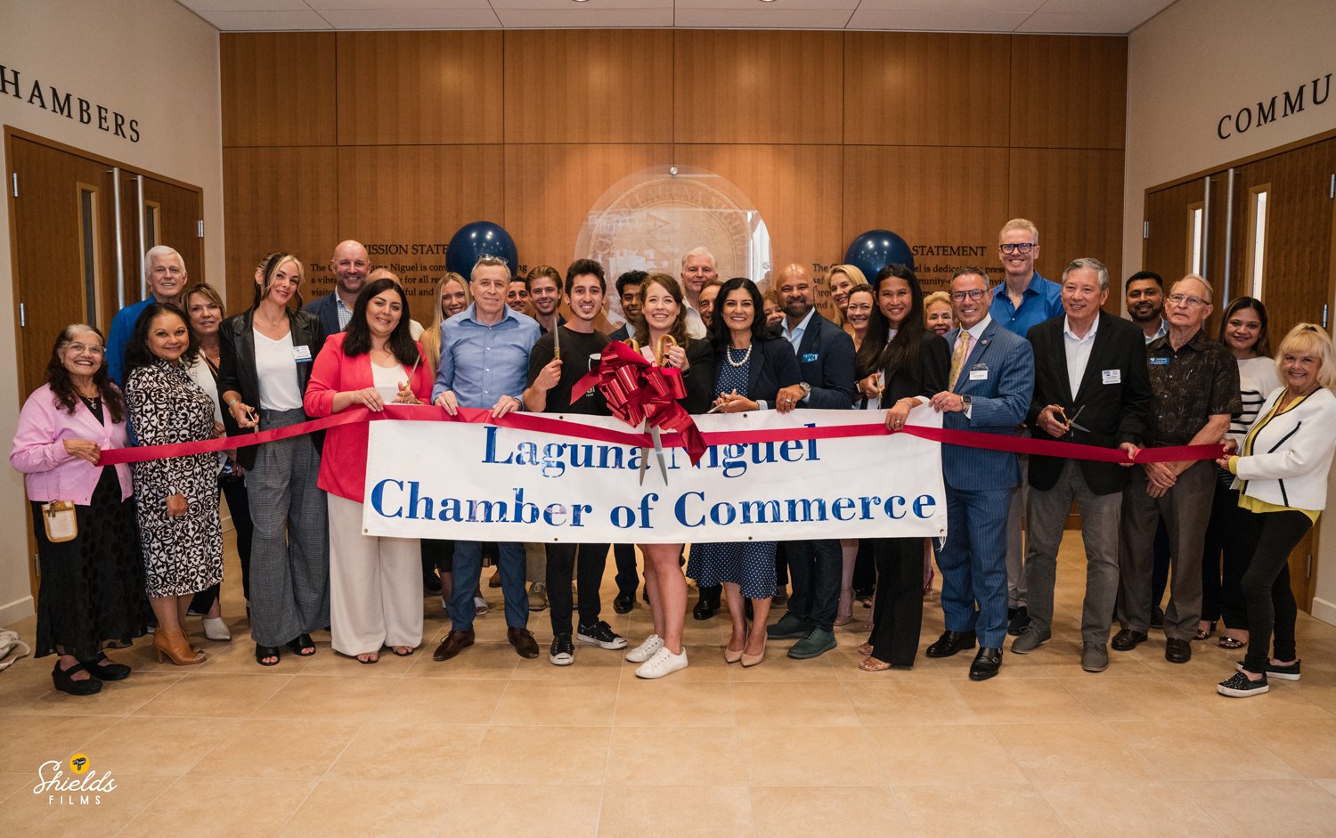 Group of people at a ribbon-cutting ceremony for the Laguna Niguel Chamber of Commerce, holding a large ribbon with a red bow, in a room with wood-paneled walls.