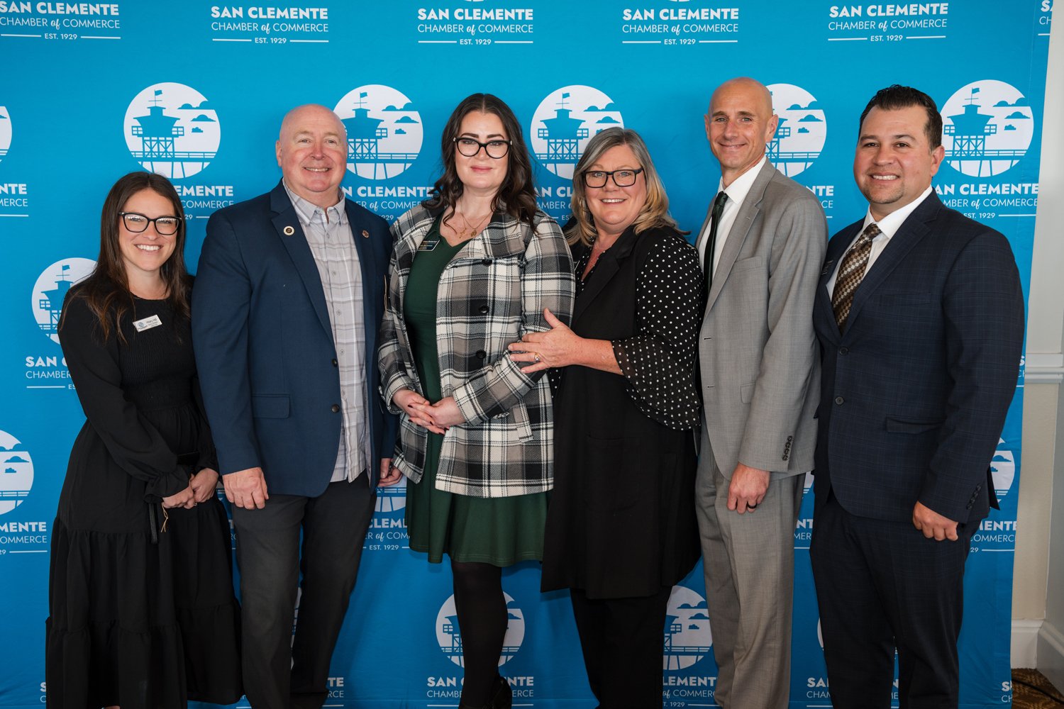 Group of seven people standing in front of a blue backdrop with the San Clemente Chamber of Commerce logo, smiling for the camera, at a professional event.