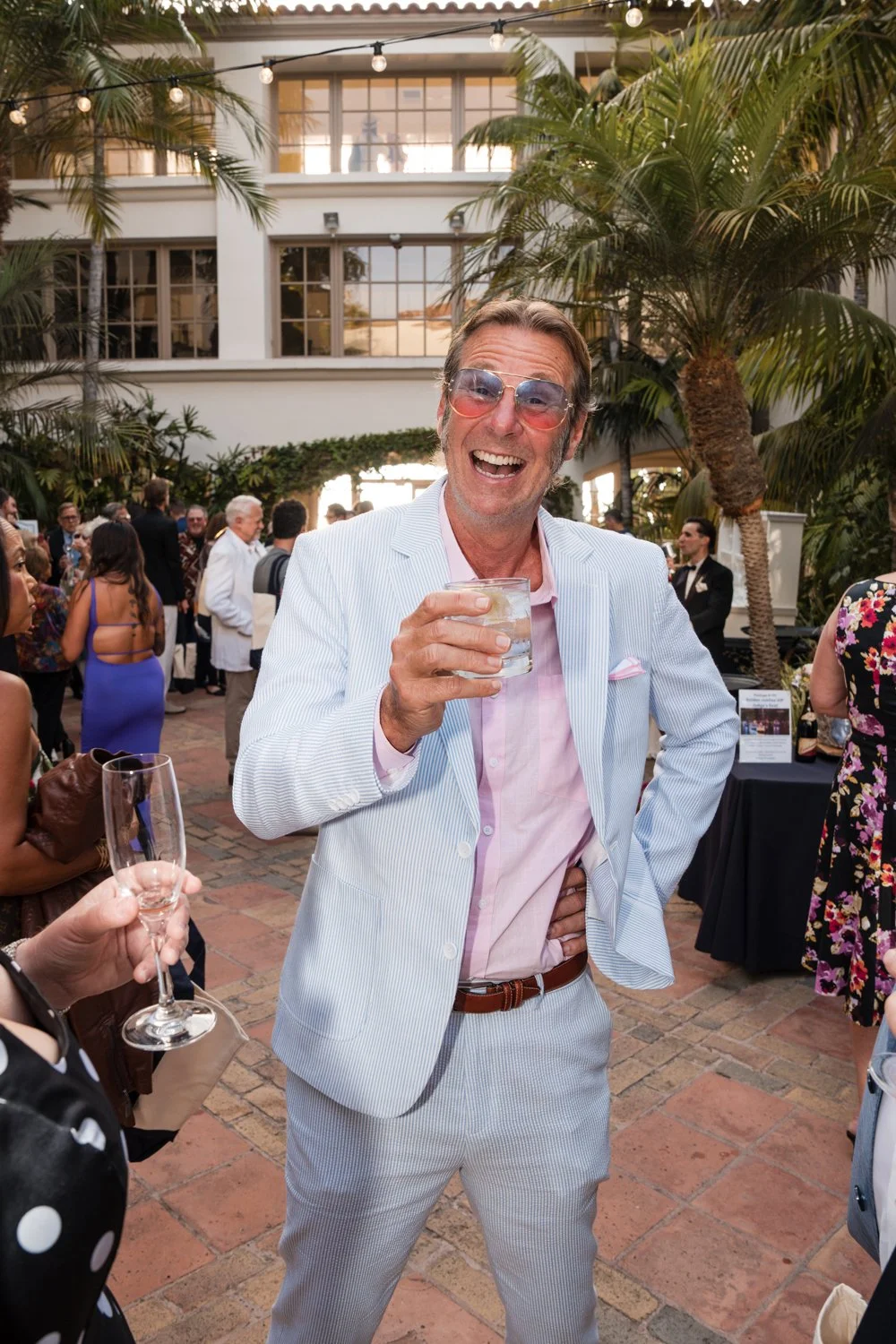 A man in a light blue and white suit holding a drink and smiling at a social gathering, with several people behind him amidst palm trees and an indoor/outdoor space with string lights.
