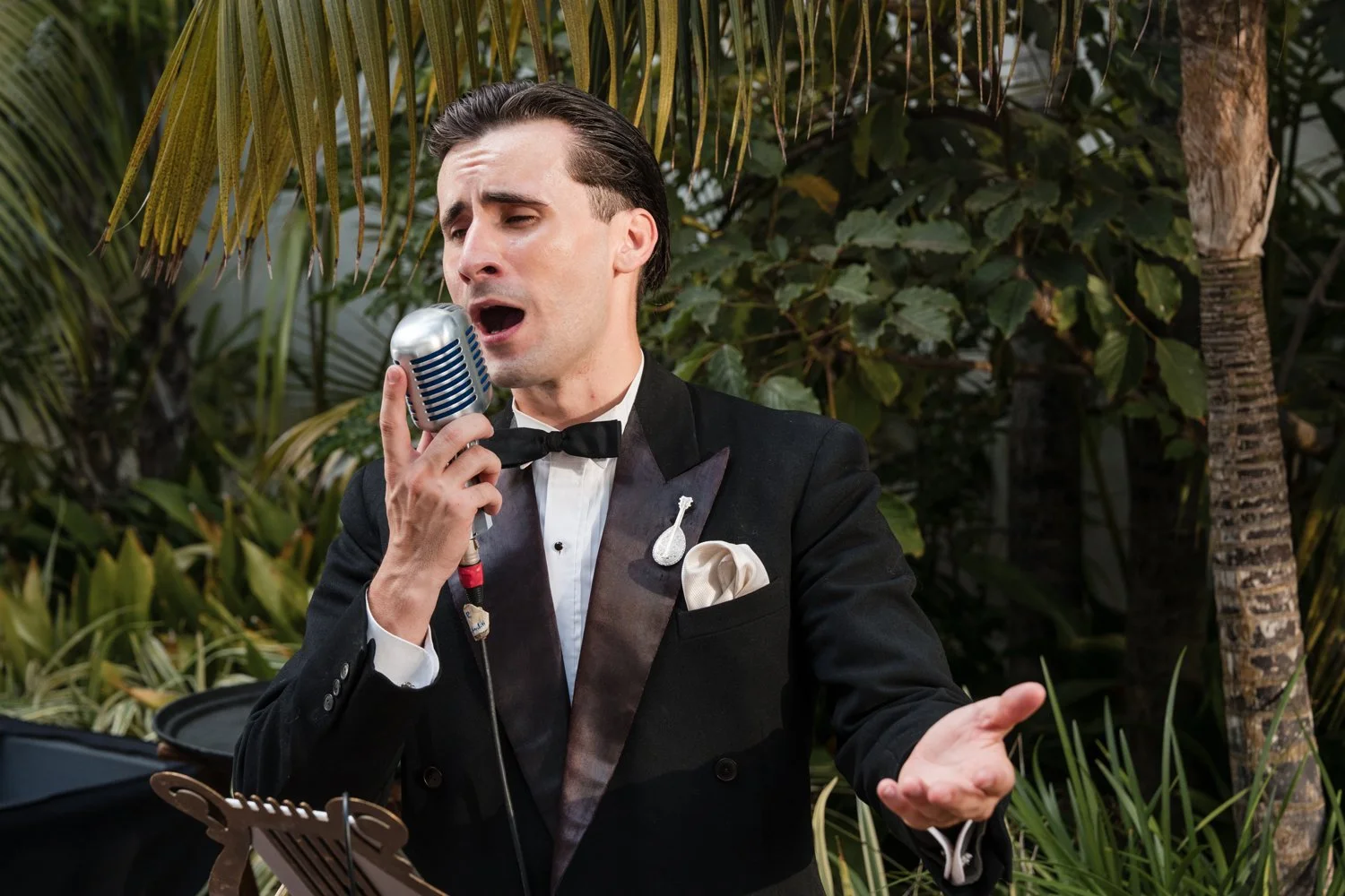 A man dressed in a tuxedo singing into a vintage microphone outdoors with dense green foliage in the background.