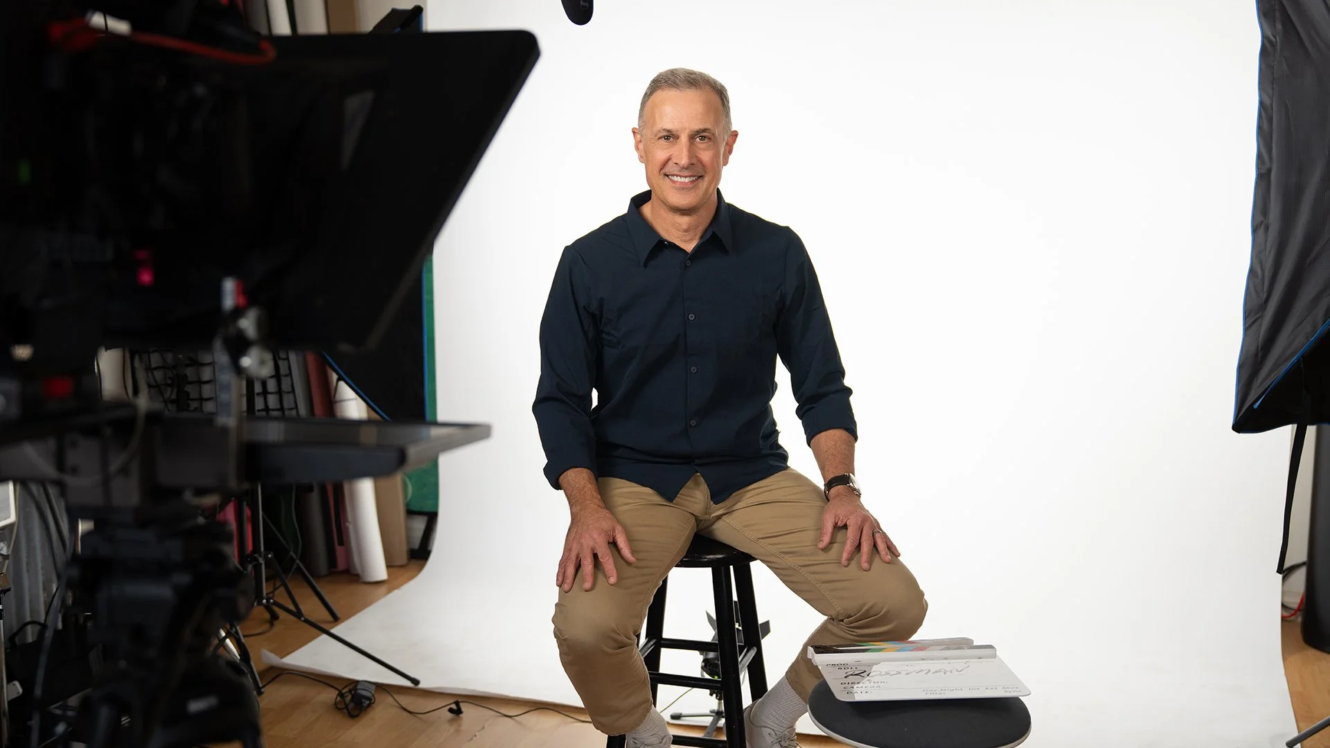 A middle-aged man with gray hair smiling, seated on a black stool in a photography studio with white backdrop, surrounded by professional lighting and camera equipment.