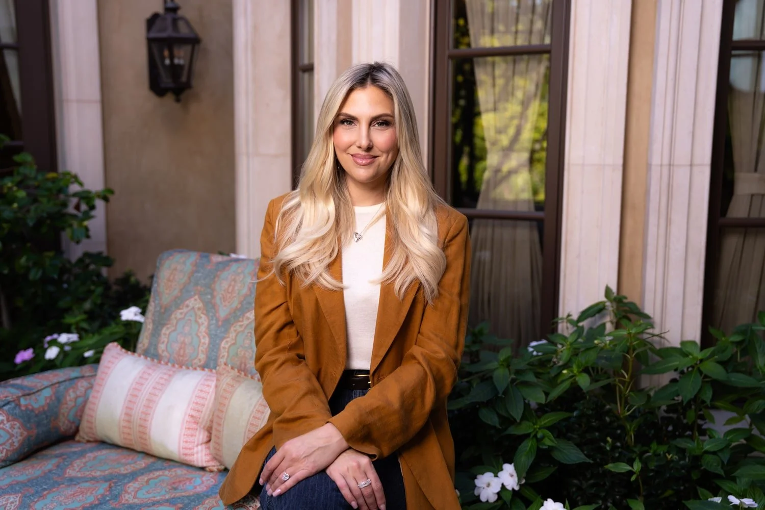 A woman with long blonde hair smiling while sitting on a patterned outdoor sofa with pillows, in front of a house with large windows and lush greenery.