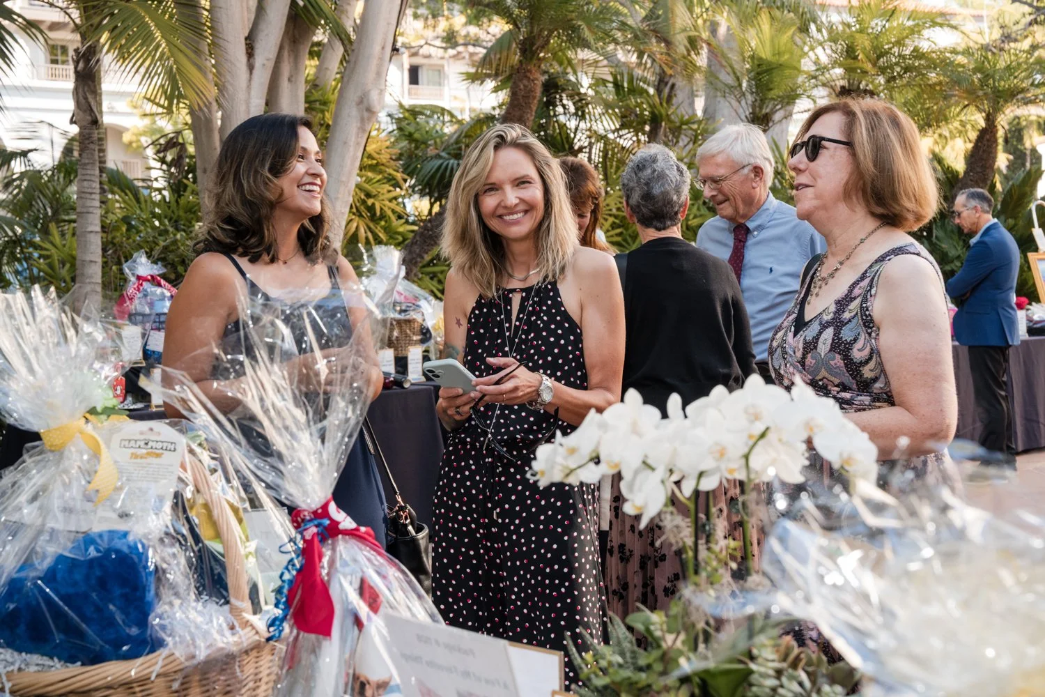 Group of women at an outdoor event, chatting and smiling, with gift baskets and flowers on tables in the foreground, surrounded by trees and greenery.