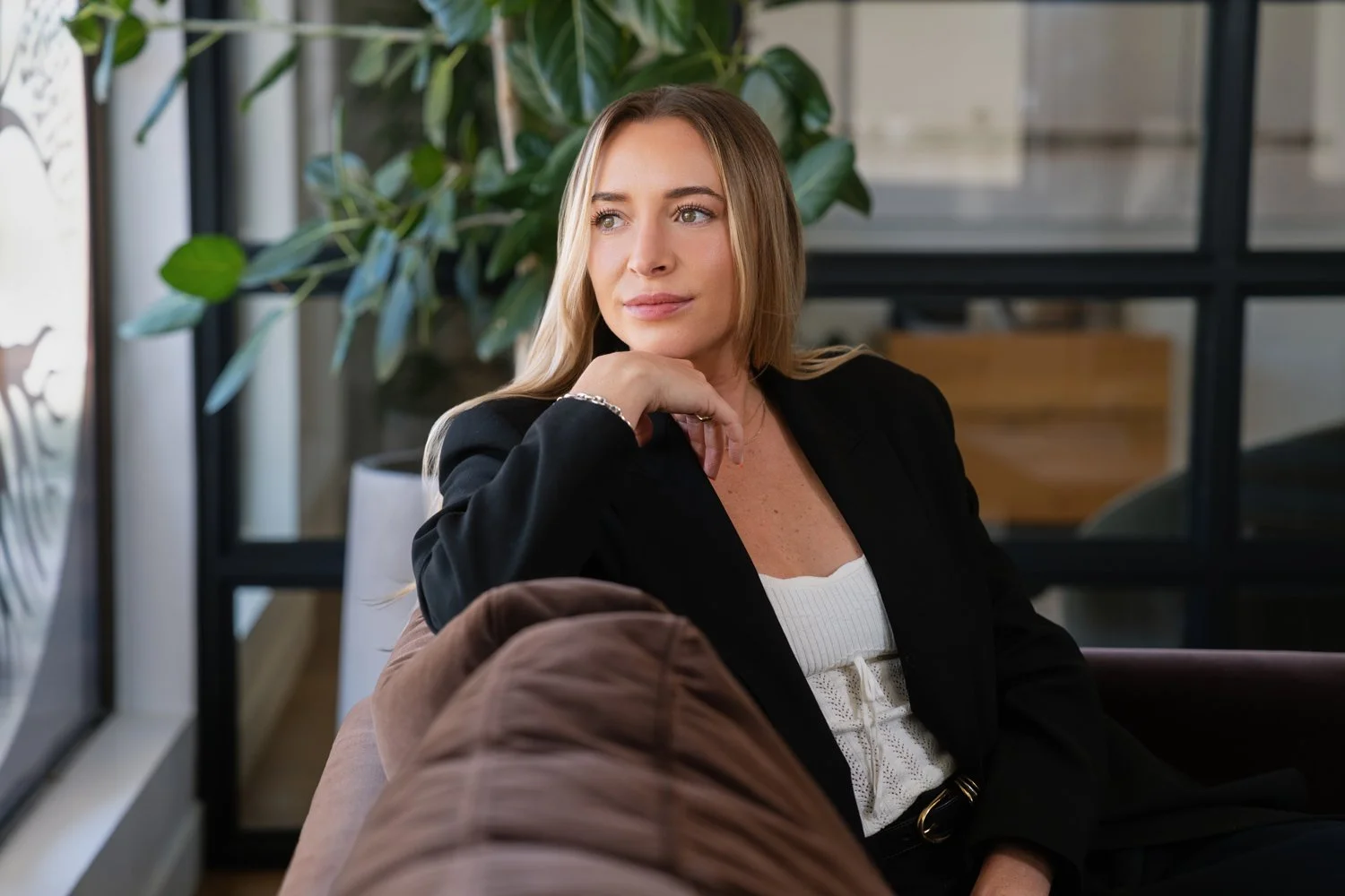 A woman with long blonde hair wearing a black blazer and white top sitting on a sofa with a plant and glass window in the background.