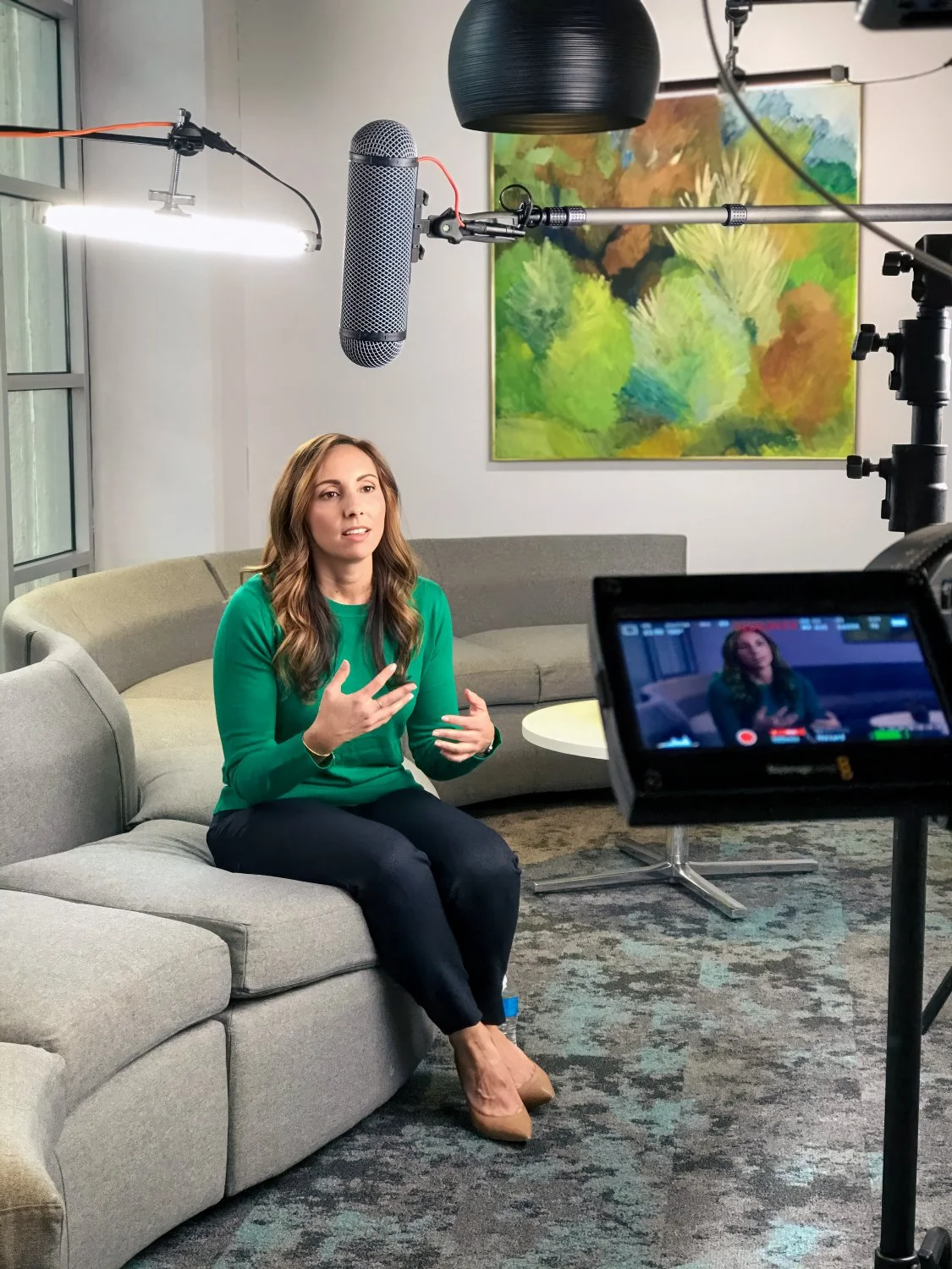 A woman in a green shirt and black pants is sitting on a gray couch, speaking during an interview or recording session, with professional video and audio equipment around her, in a room with a colorful abstract painting on the wall.