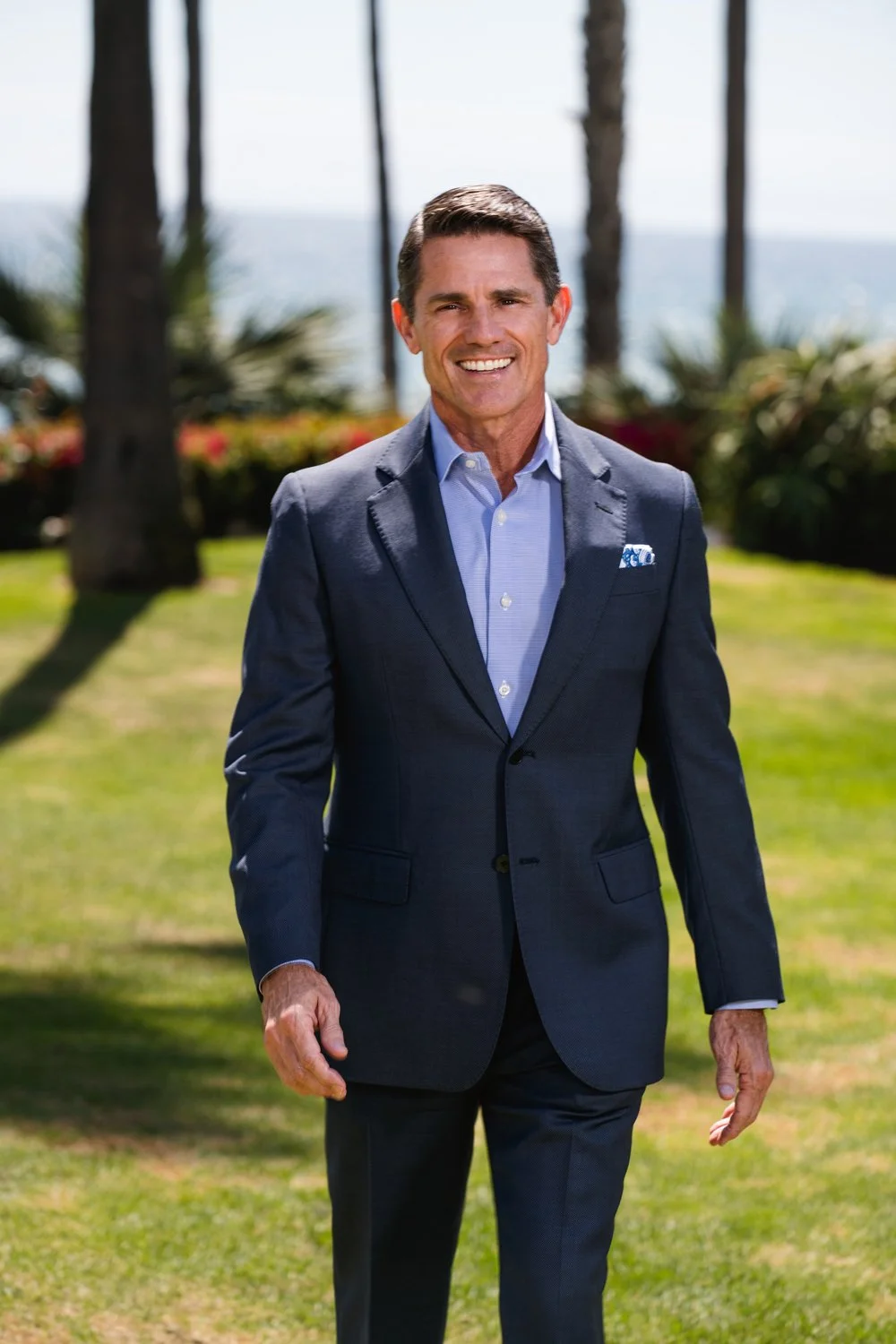 A man in a dark blue suit and light blue shirt standing outdoors on a grassy area with trees and the ocean in the background, smiling at the camera.