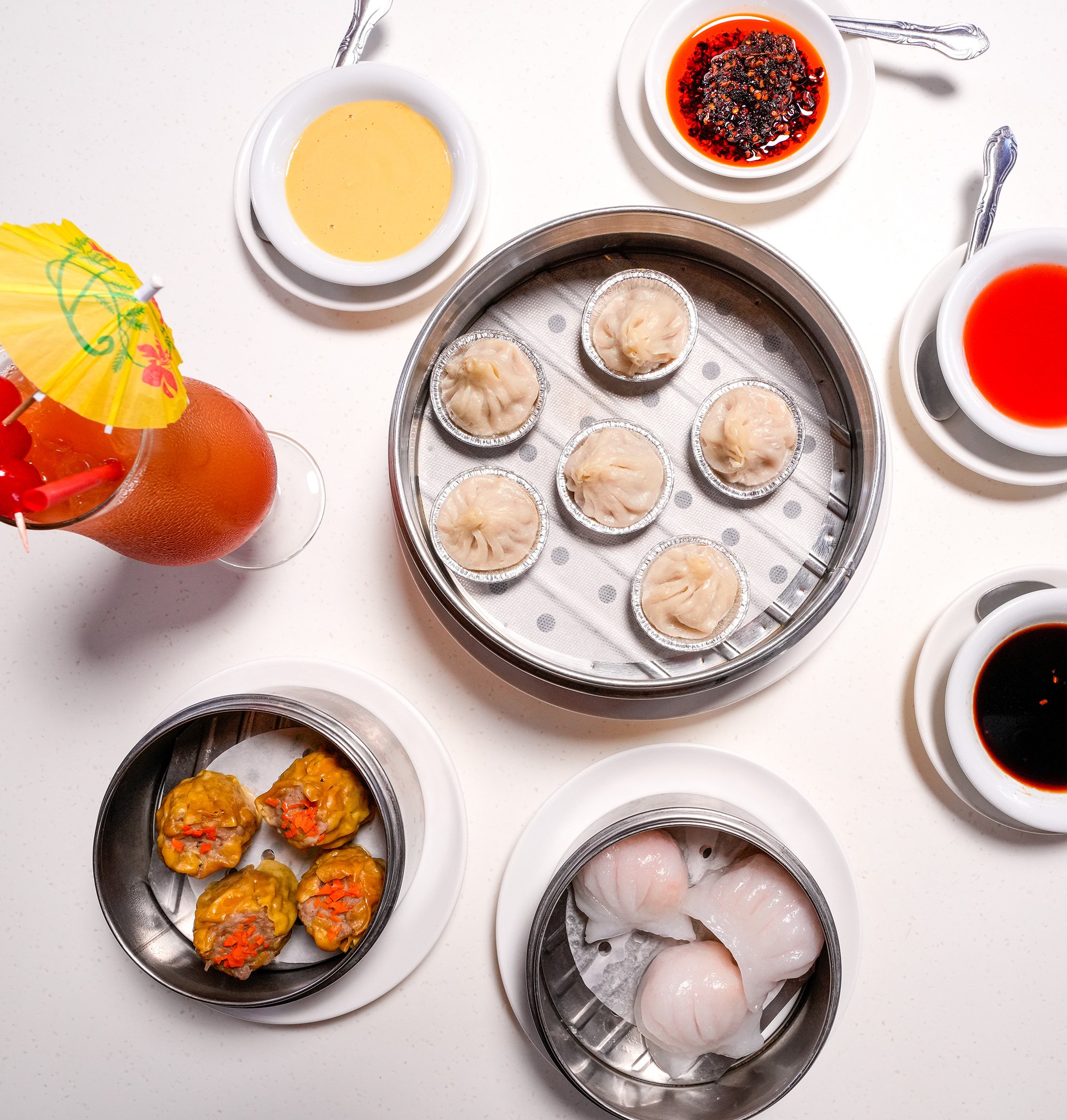 Variety of dishes including steamed dumplings, side sauces, and a drink with a small paper umbrella on a white table.