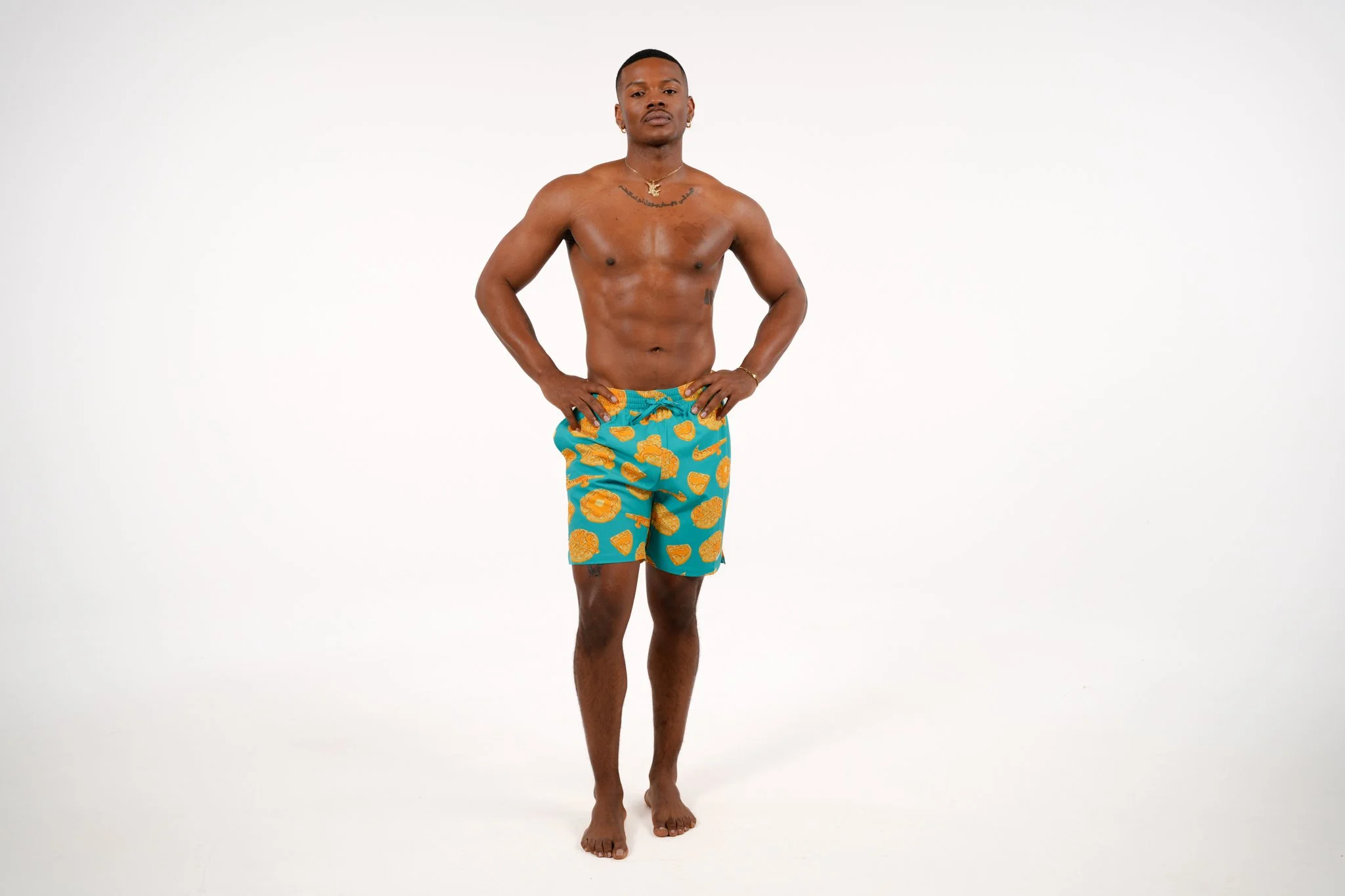 A shirtless man wearing blue shorts with orange pineapple patterns, standing against a plain white background.