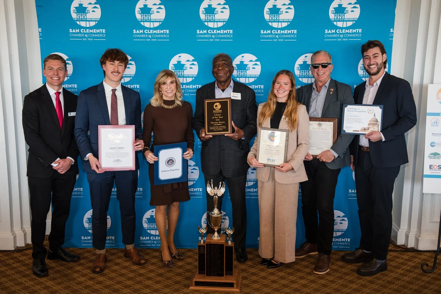 Group of eight people standing in front of a blue backdrop that says 'San Clemente Chamber of Commerce' with a picture of an oil rig. They are holding certificates and plaques, with a large trophy in front of them.