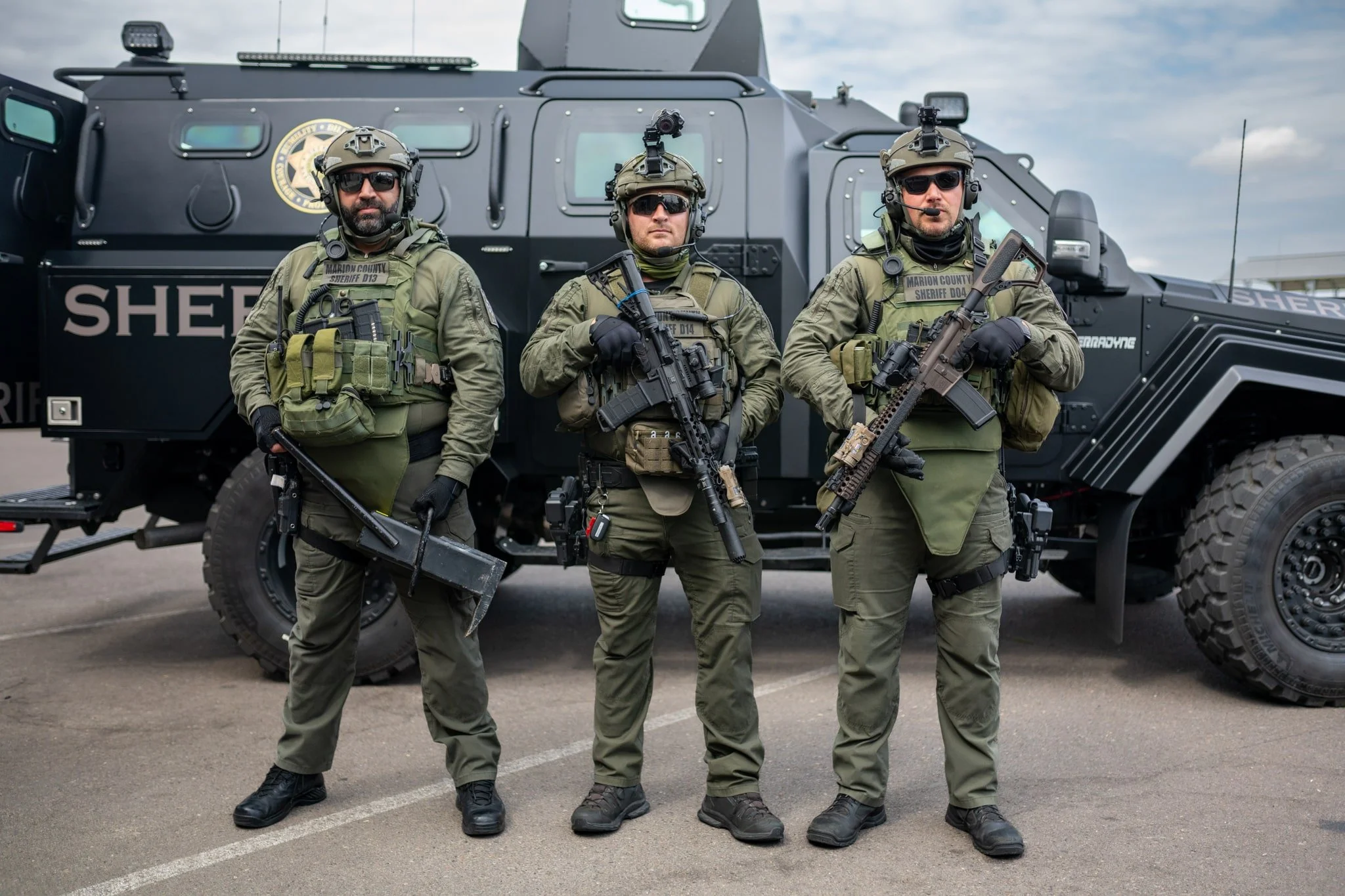 Three police officers in tactical gear and sunglasses stand in front of a large armored vehicle marked 'Sheriff'.