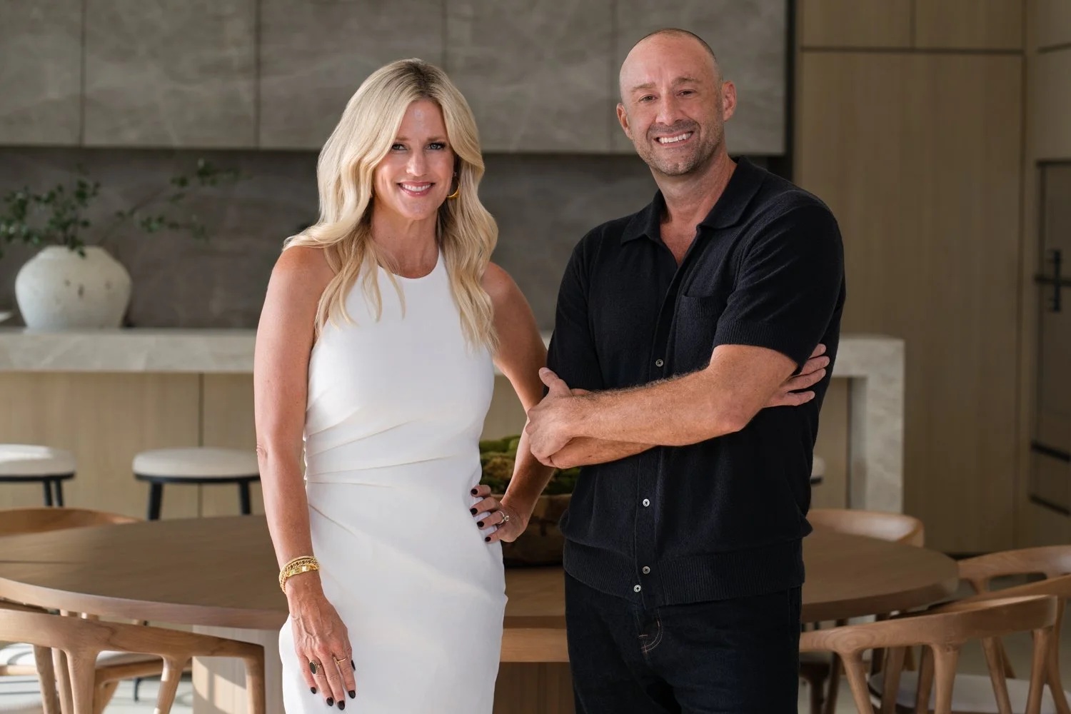 A woman with long blonde hair and a man with a shaved head, both smiling and standing in a modern kitchen. The woman is wearing a white sleeveless dress with jewelry, and the man is dressed in a black polo shirt with crossed arms.