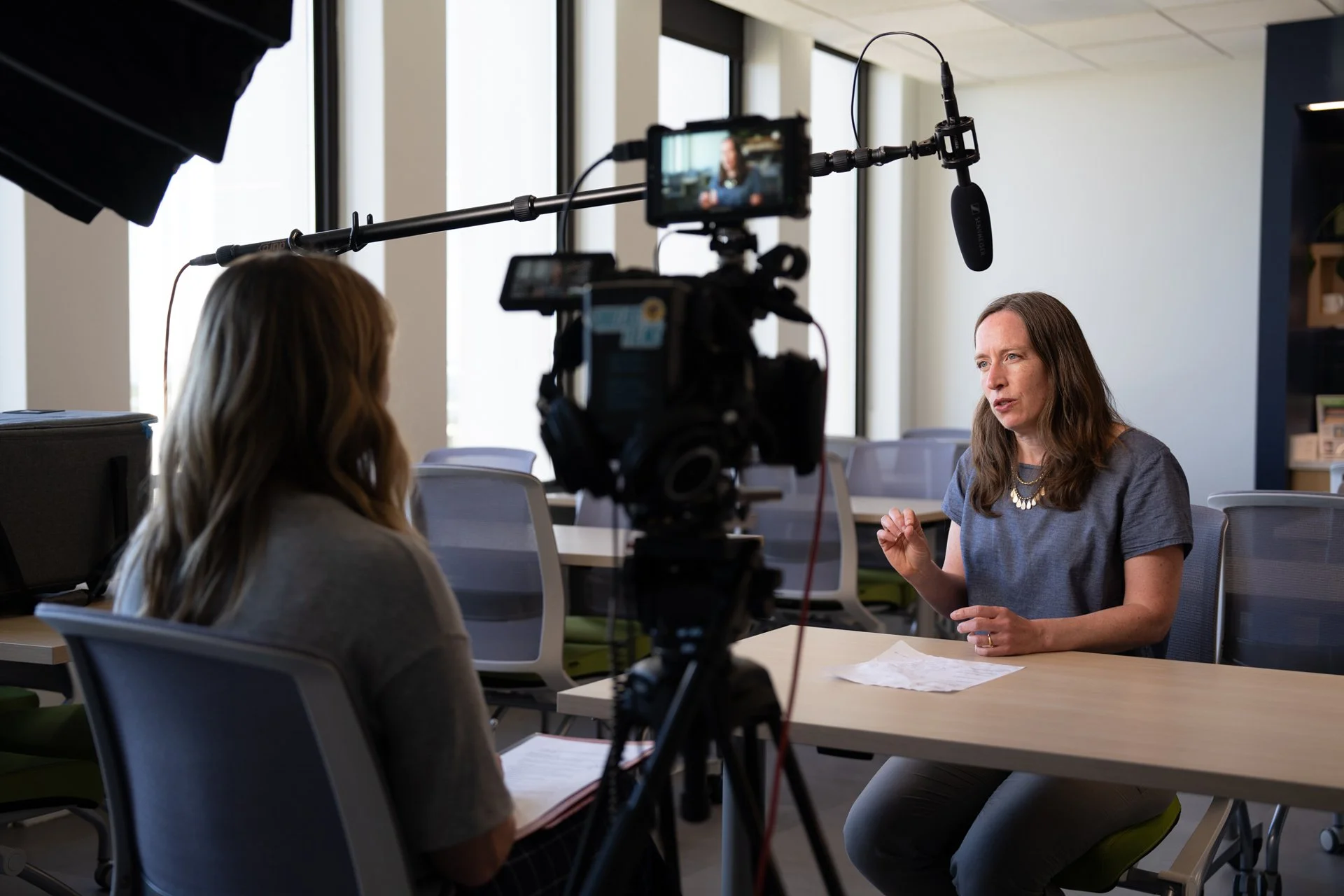 A woman being interviewed in an indoor office setting with blinds on the window in the background. A professional camera with a microphone is recording her, and a person with long hair is operating the camera. The woman appears to be speaking and gesturing with her hand while sitting at a desk.