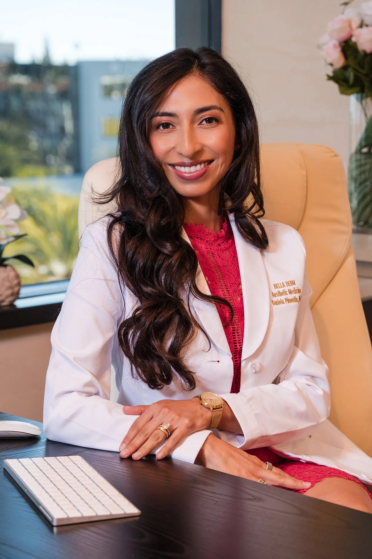 A woman with long dark hair and a warm smile sitting at a desk in an office, wearing a white coat with embroidered text and a red lace top underneath. She's resting her hands on the desk, which has a keyboard and mouse, and is wearing a gold watch an