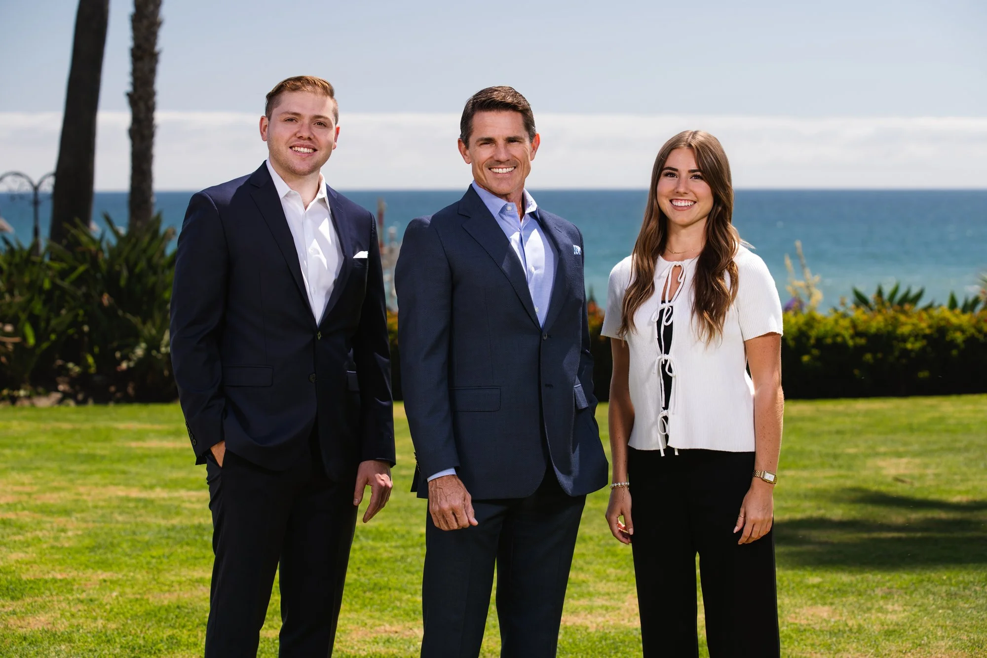 Three professionally dressed people, two men and a woman, standing outdoors near the coast with a view of the ocean and palm trees in the background, smiling at the camera.