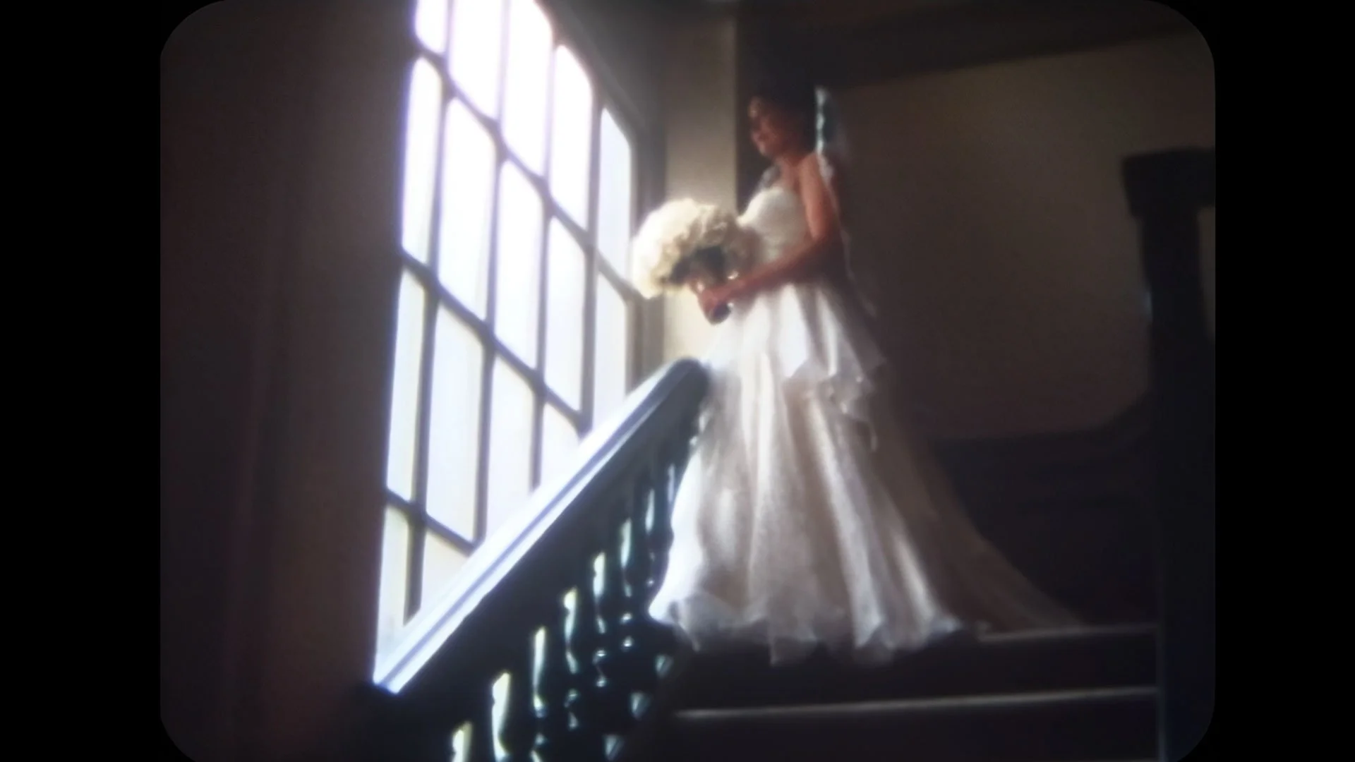 A bride in a white wedding gown with a bouquet standing at the top of a staircase near a large window.