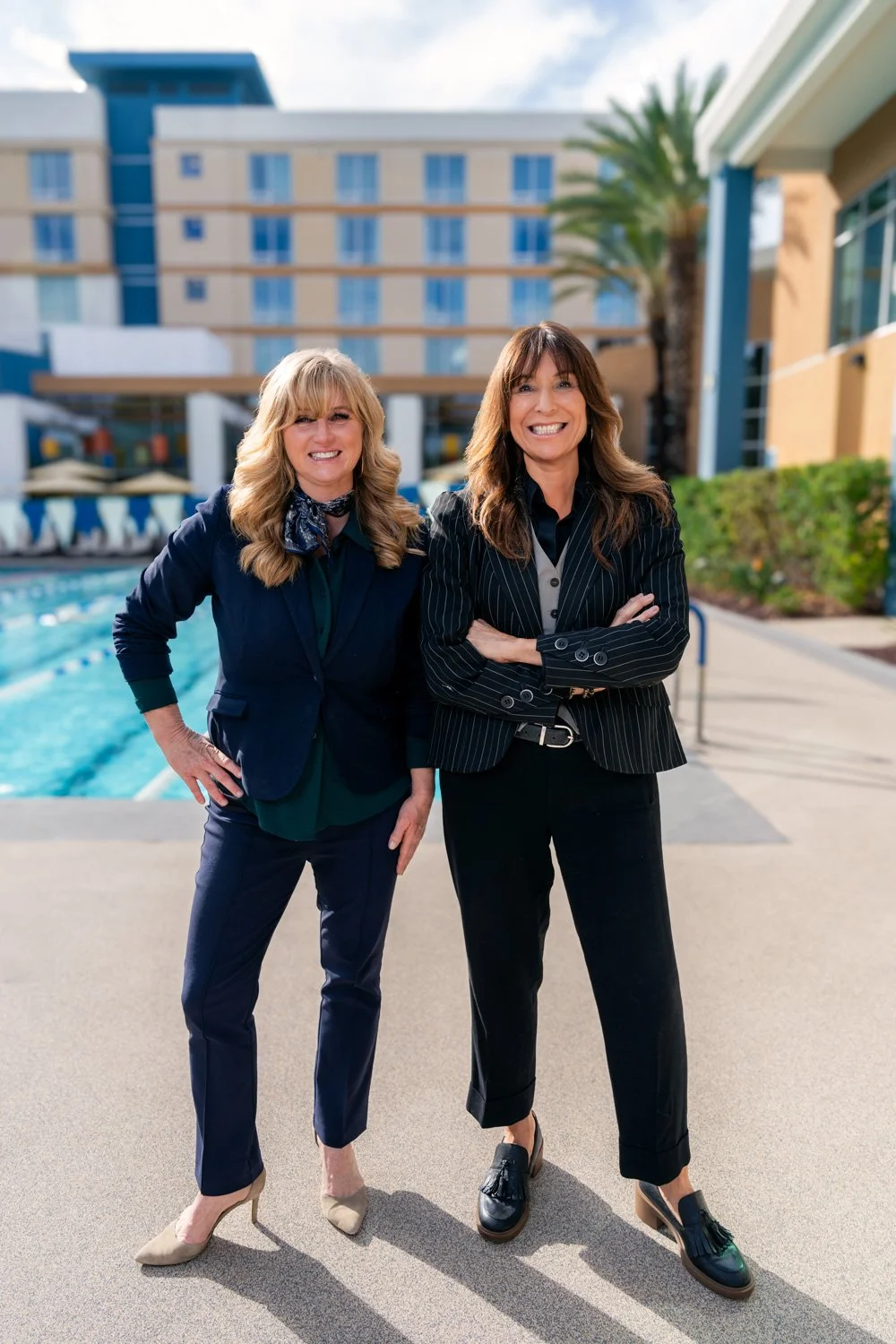 Two women in business attire standing outdoors by a swimming pool at a hotel or resort with a tall building in the background, smiling at the camera.