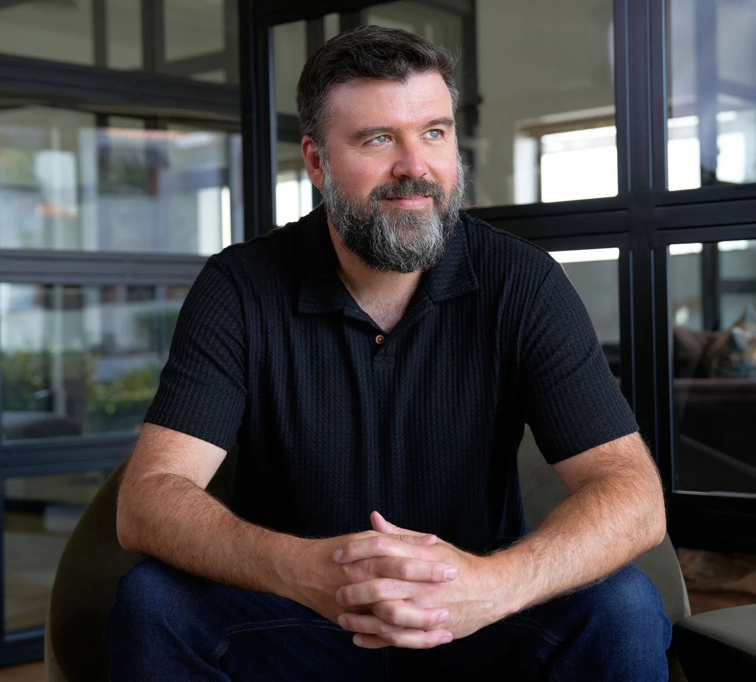 A man with short dark hair, a beard, wearing a black collared shirt, sitting indoors with hands folded, looking to the side.