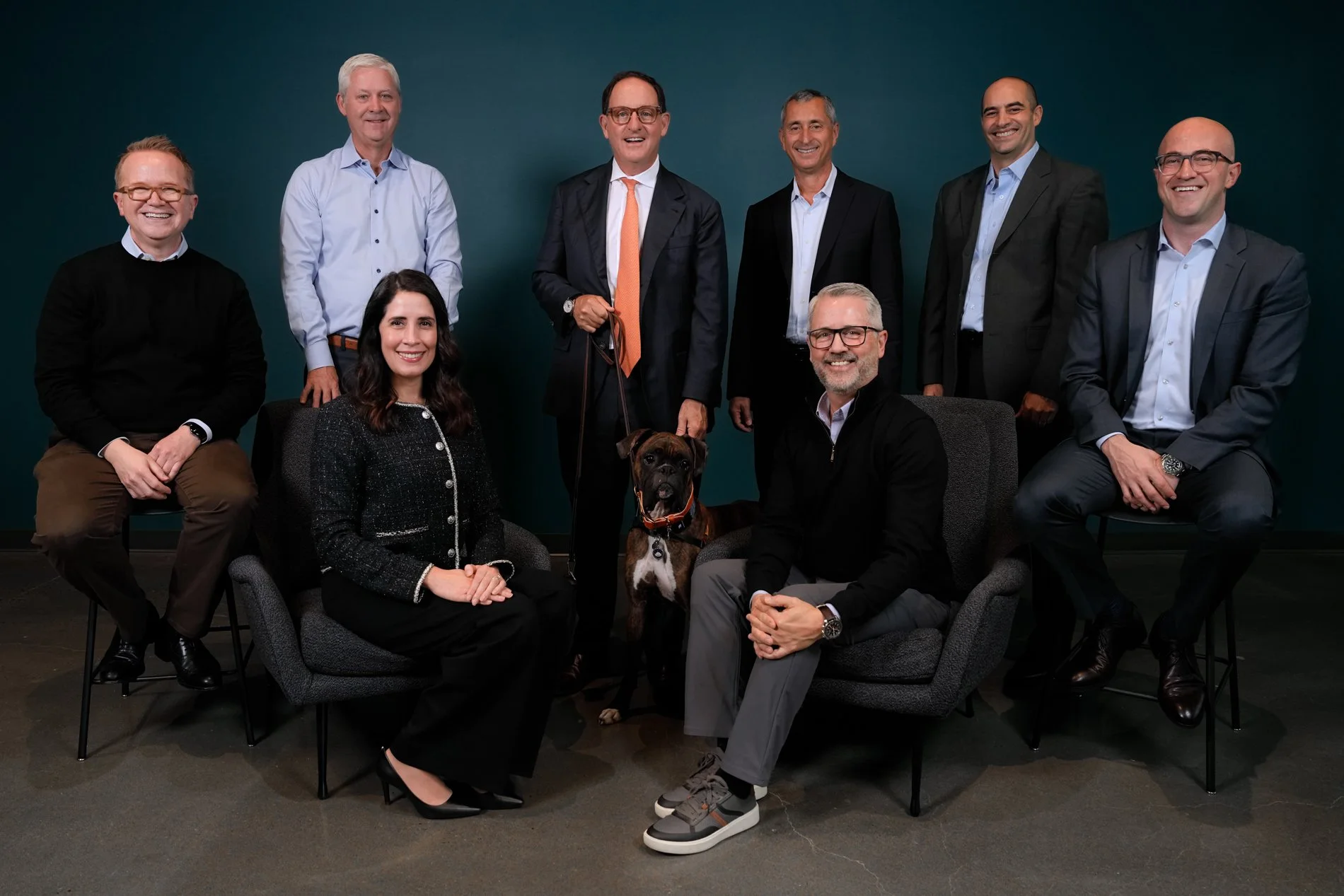 A group of nine diverse professionals, including two women and seven men, posing for a photo in an office setting. They are dressed in business attire, with some seated and others standing. There are two dogs in the image, one being held on a leash b